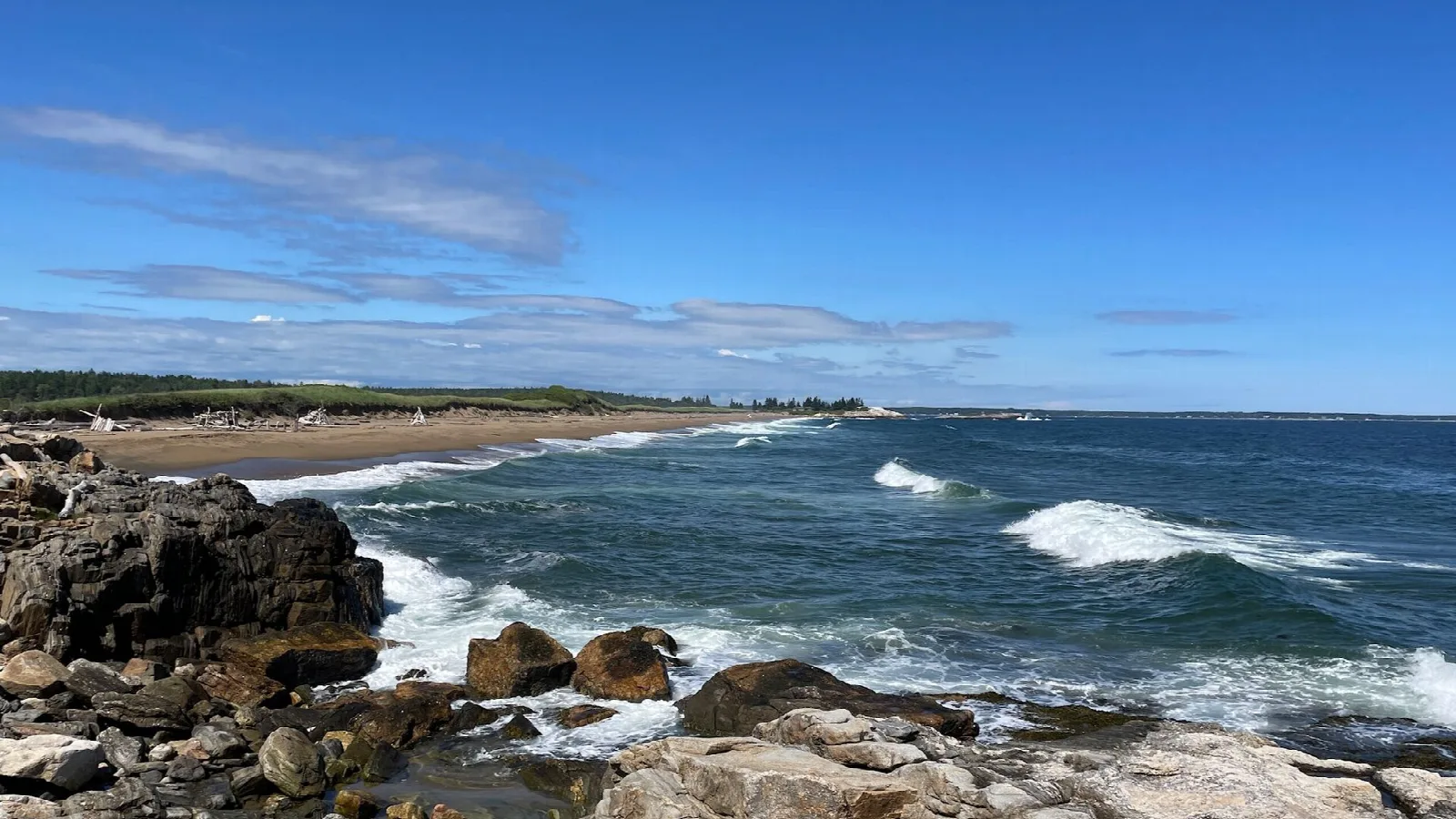 Surf rolling onto a rocky beach with a long stretch of sand beyond, under blue sky