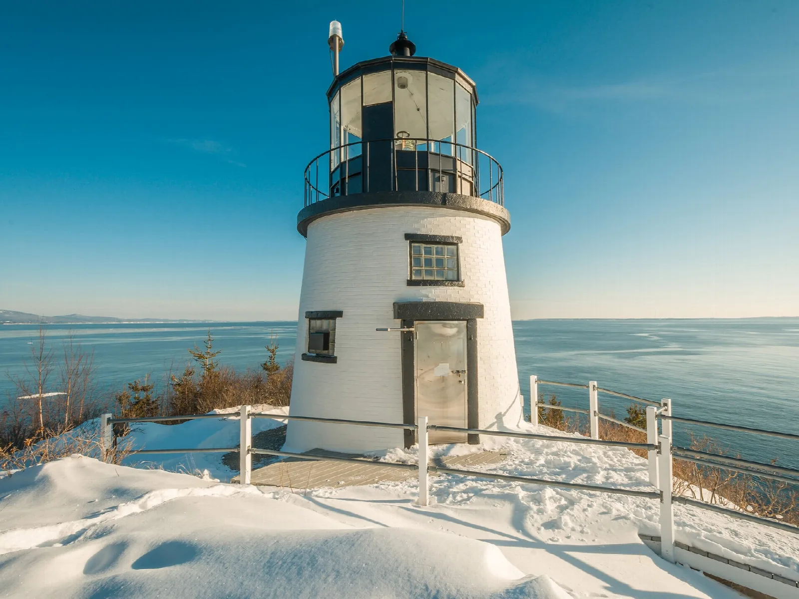 White lighthouse tower on a snowy clifftop above a calm blue bay