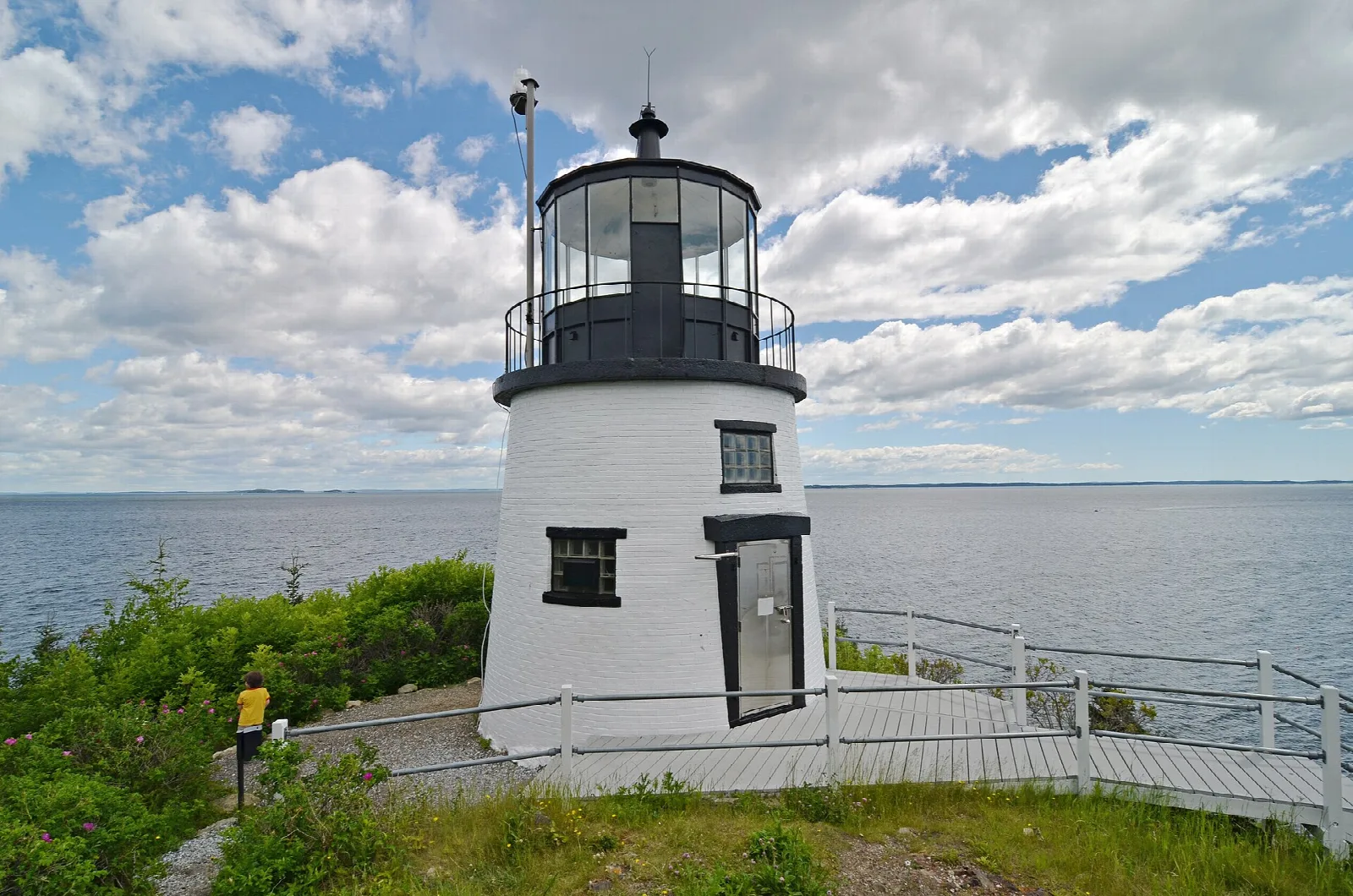 White lighthouse with black lantern on a grassy rise, wooden walkway and bay behind