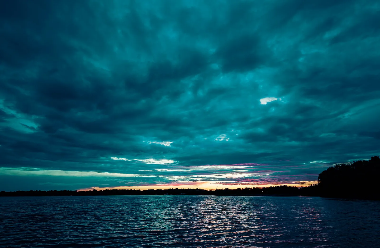 Calm lake at dusk under dark teal clouds with a wooded far shore
