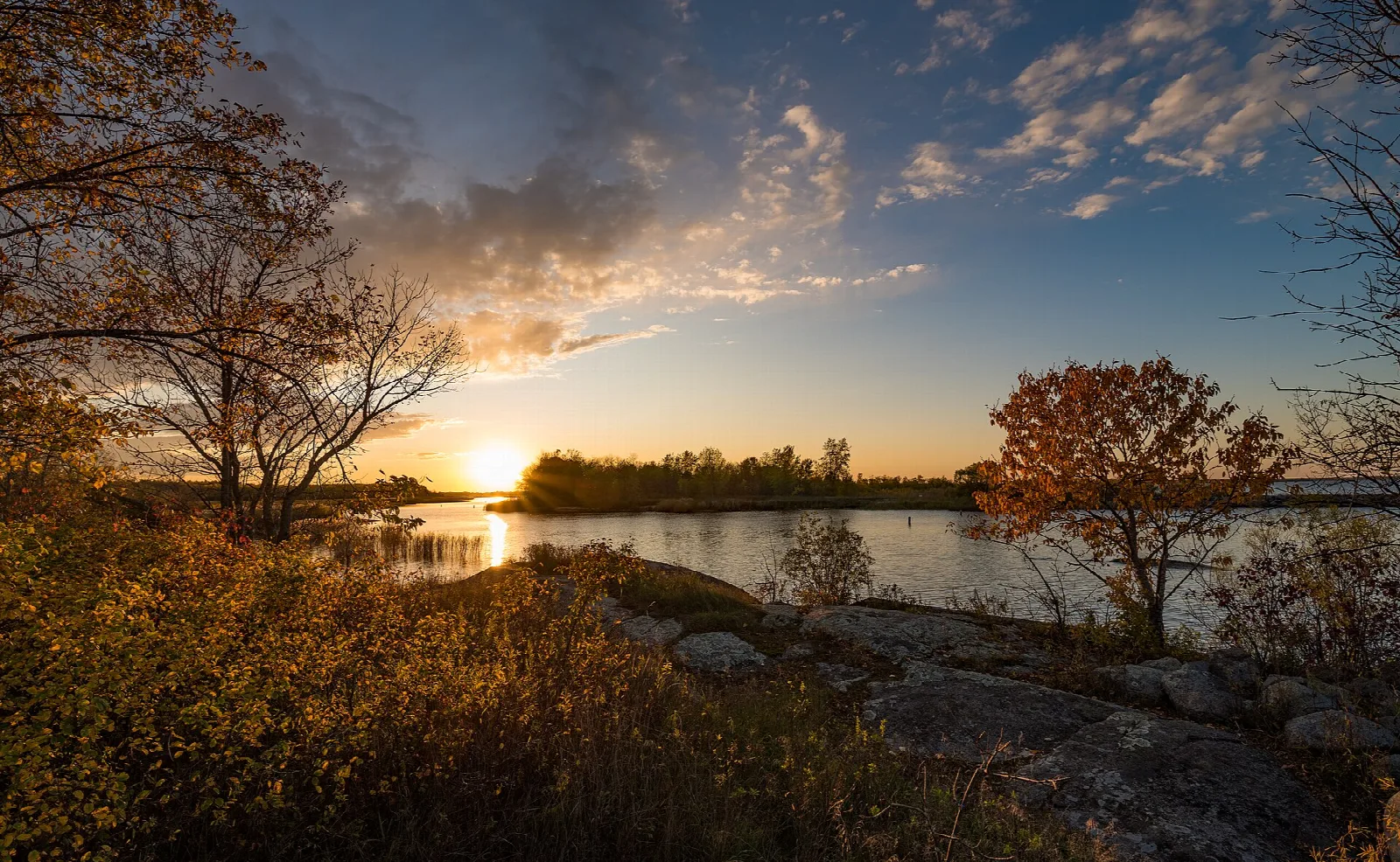 Sun setting over open water seen past rocks and autumn shrubs
