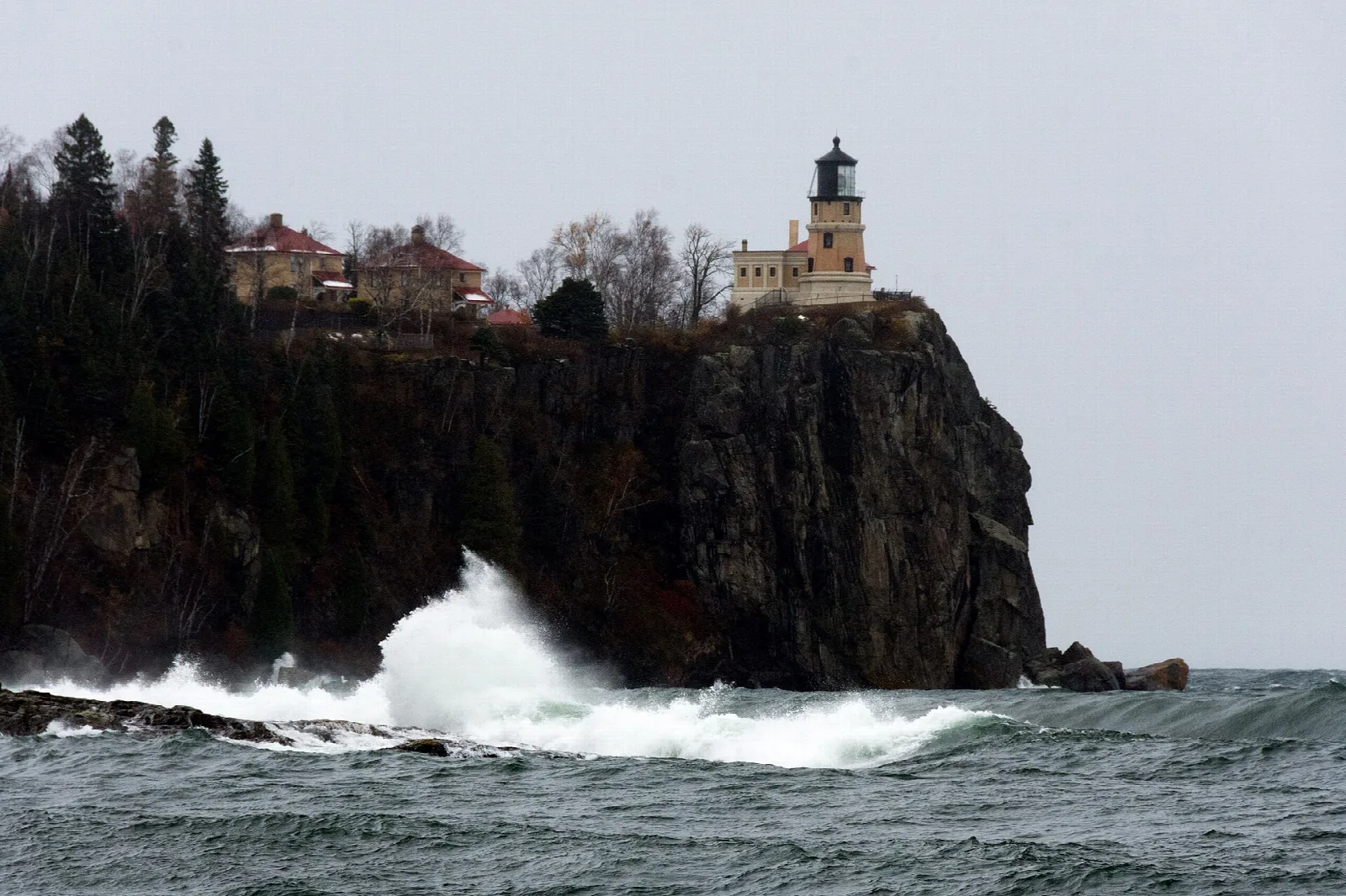 Wave crashing at the foot of a dark cliff with the lighthouse above