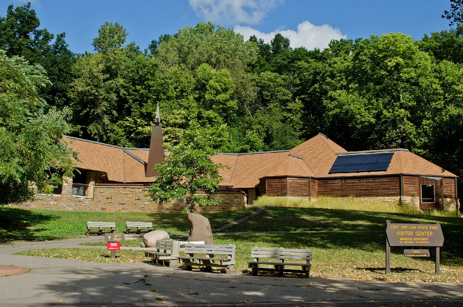 Low brown wood building with a sloped roof, benches and trees in front