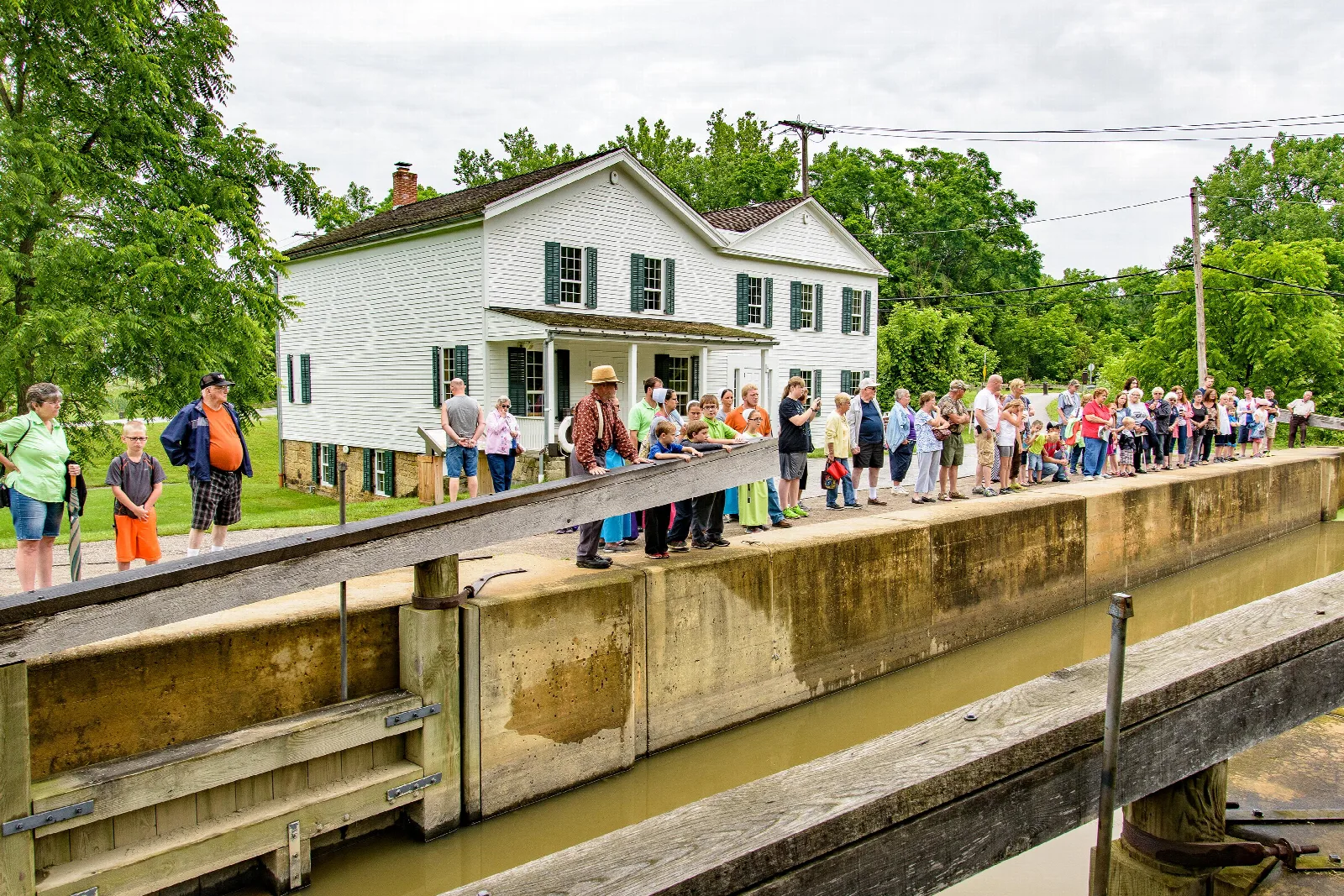 Visitors line the concrete edge of a canal lock filled with water, in front of a white building.