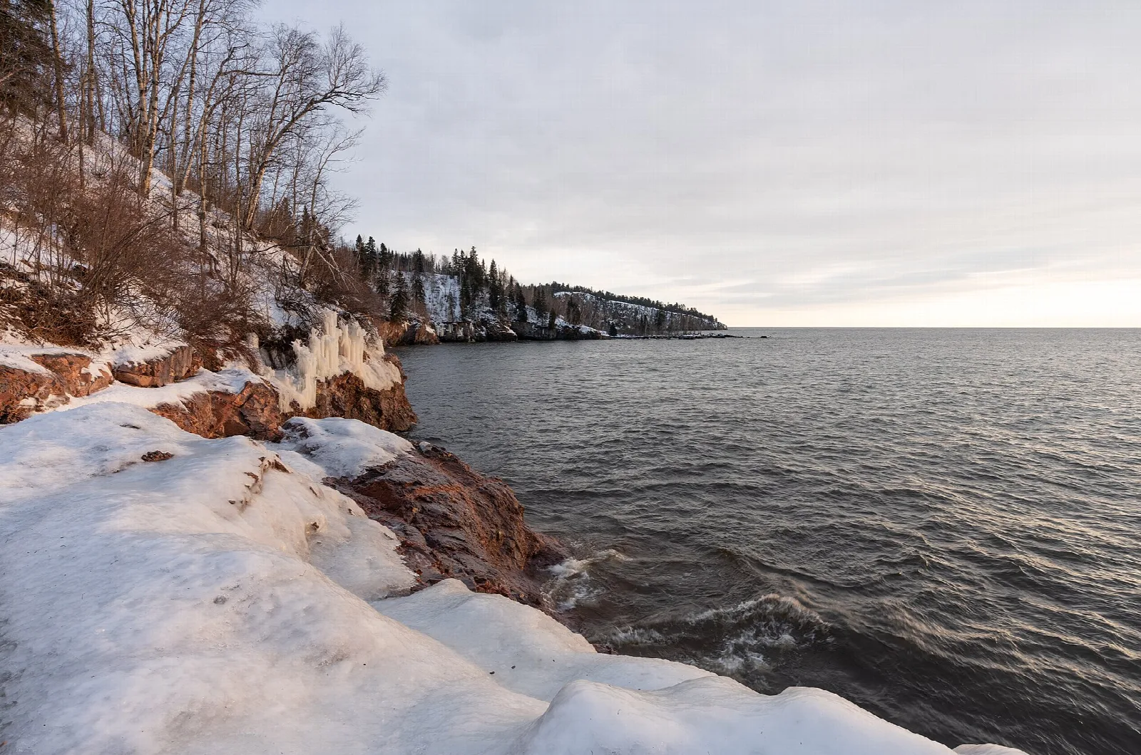 Snow-covered rocky shore along a wide lake under a gray sky
