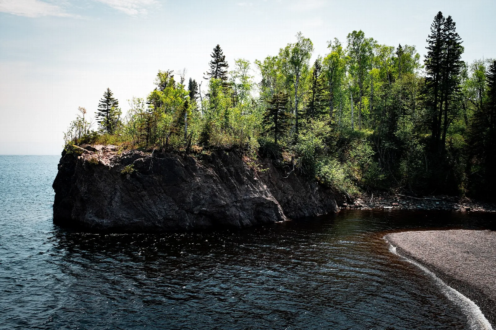 Rocky point topped with trees beside a pebble beach and open water