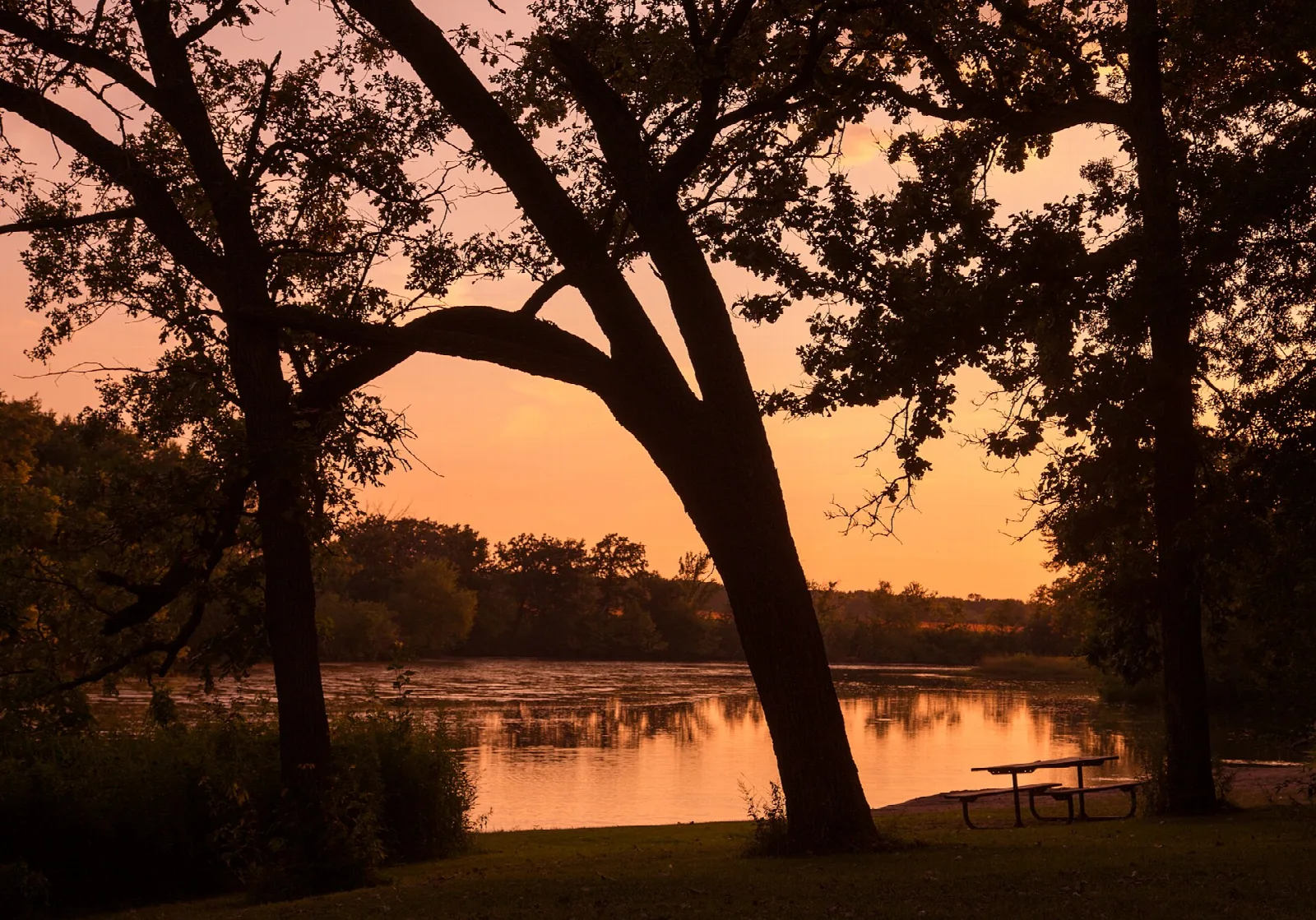 Picnic table by a lake at sunset with dark trees against an orange sky