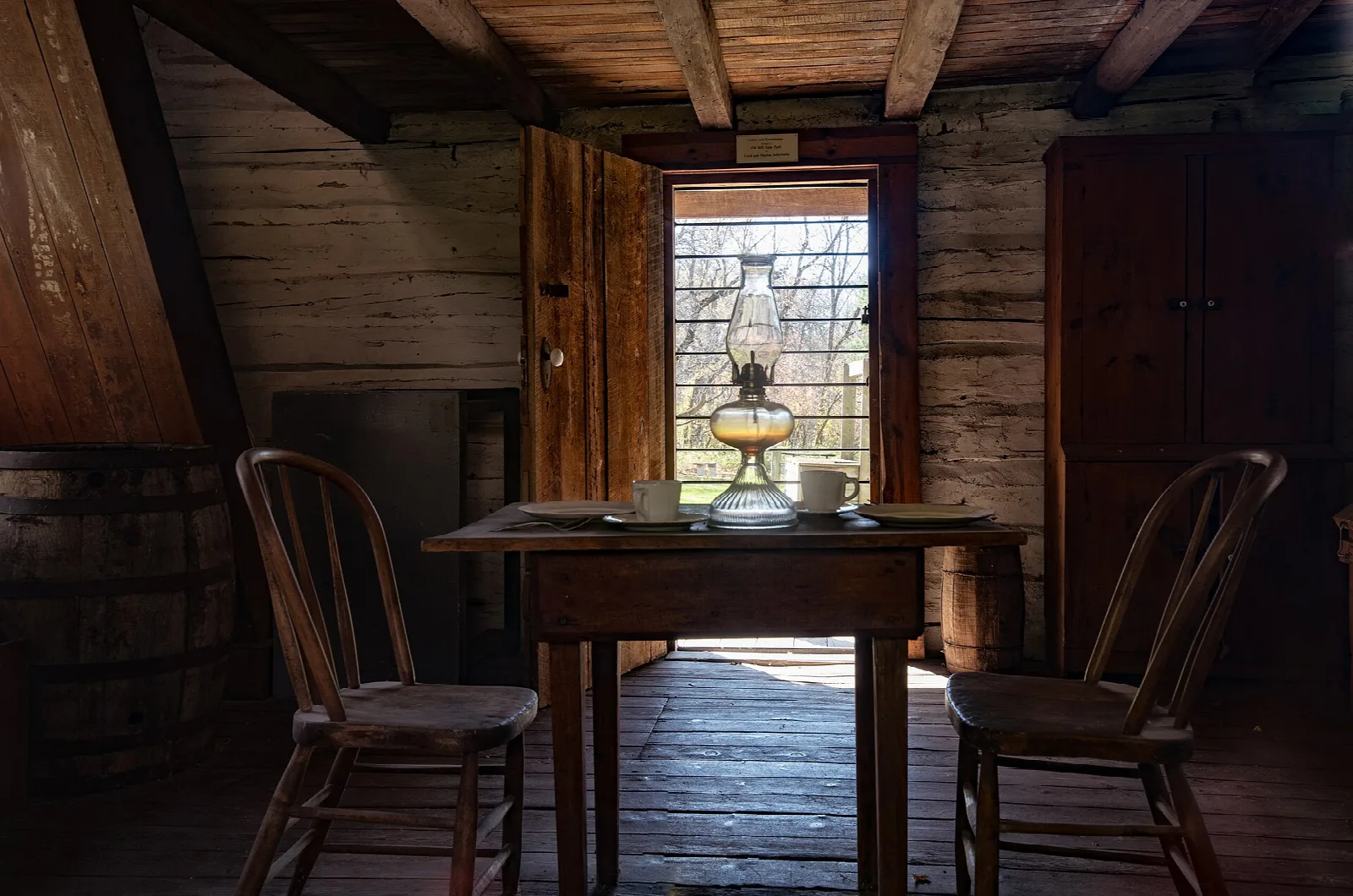 Inside a log cabin with a wooden table, two chairs and an oil lamp by the window