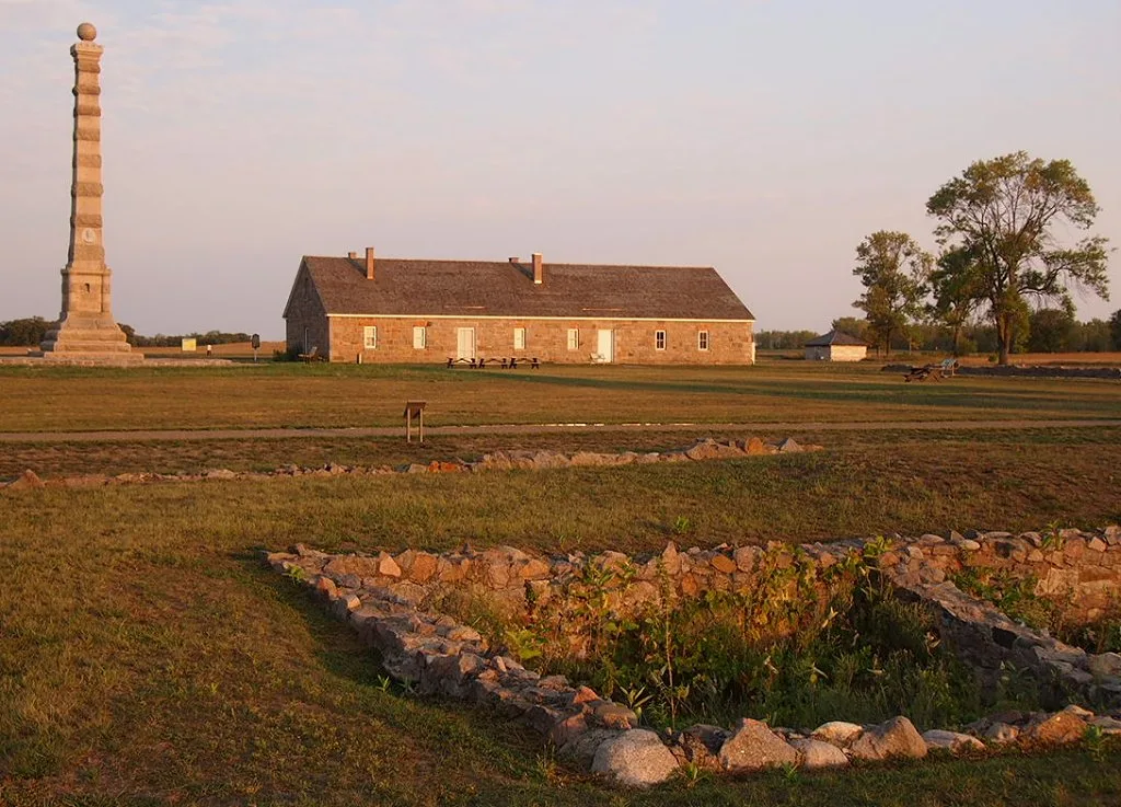 Stone barracks building and a tall monument at sunset behind a low stone foundation