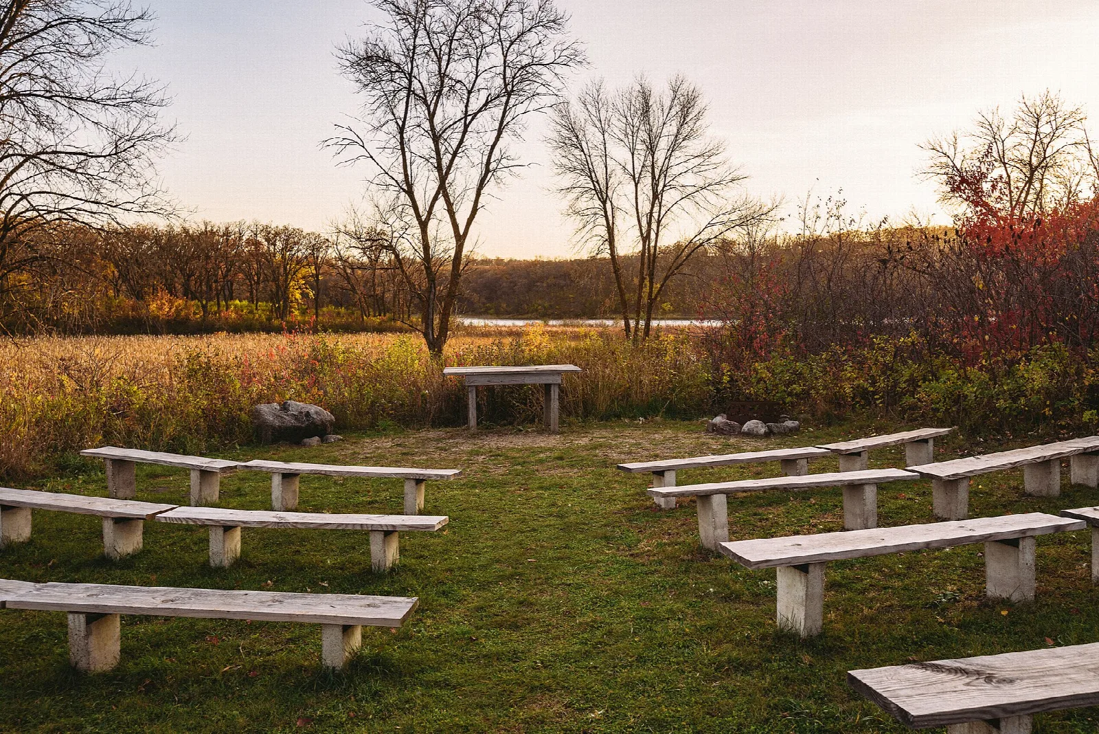Wooden benches in a circle on grass near a lake at sunset