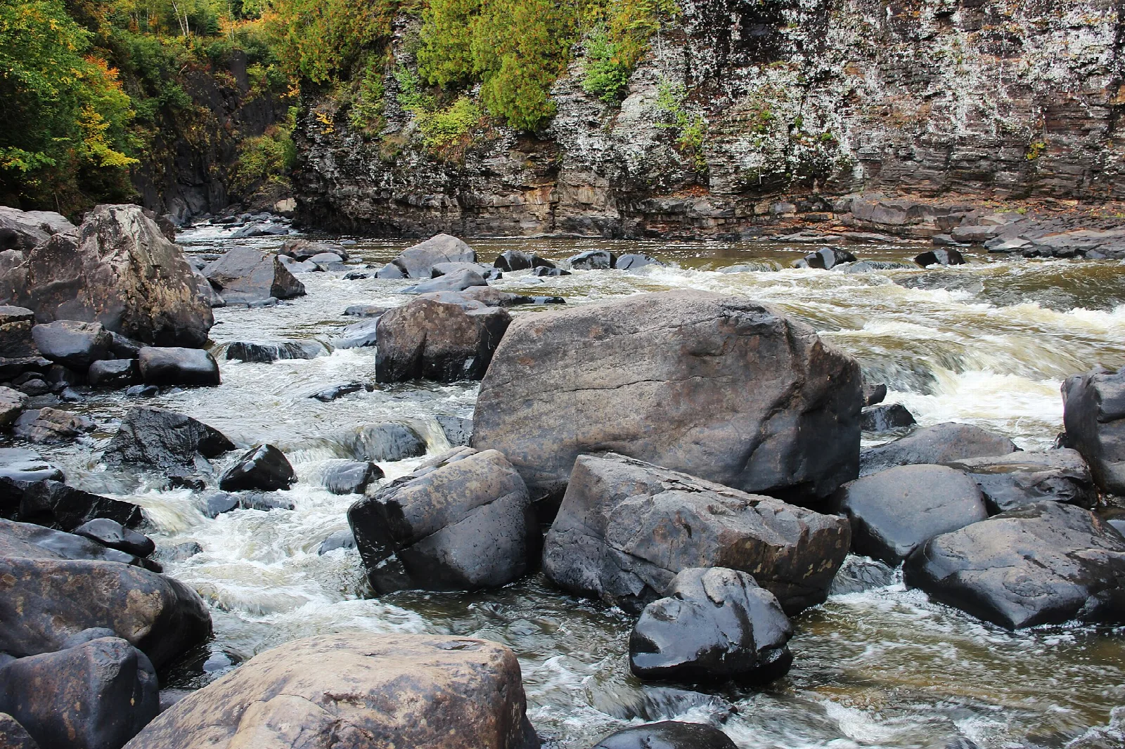 River rushing over large boulders below a rock wall