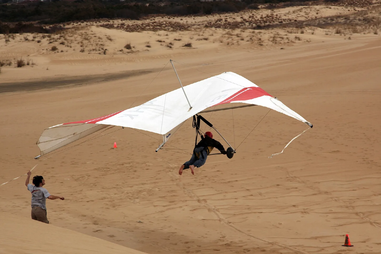 A hang glider with a white and red wing flying low over a sandy dune while a person watches