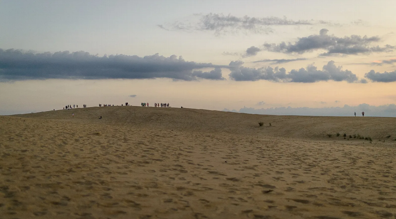Wide sandy dune at dusk with a line of people on the crest and soft clouds