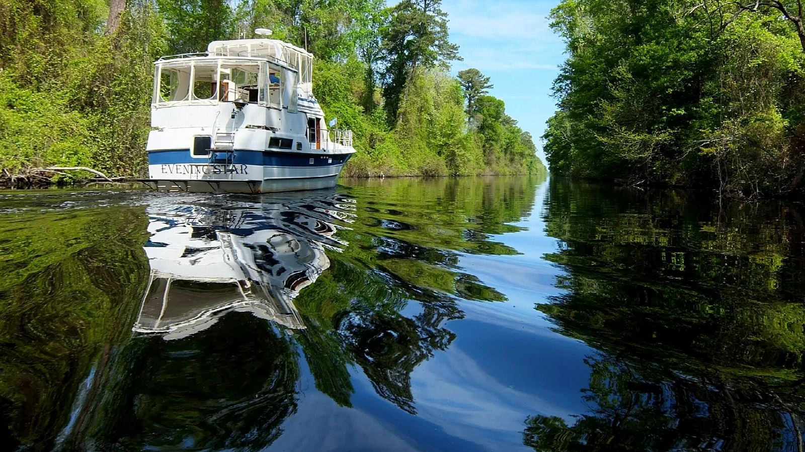 A white boat travelling down a narrow canal lined with trees