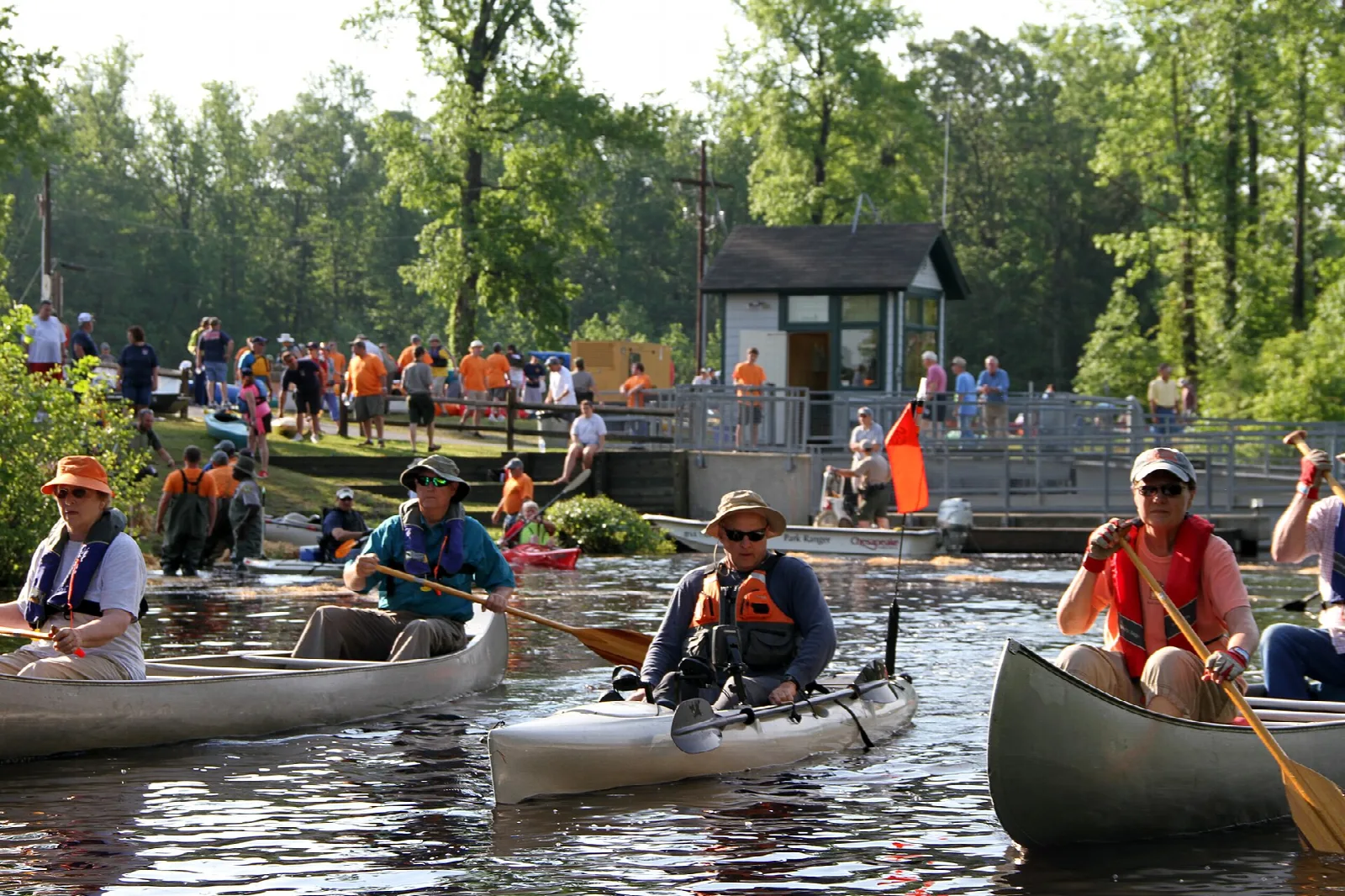 Canoes and kayaks on the water near a dock with people gathered on the bank