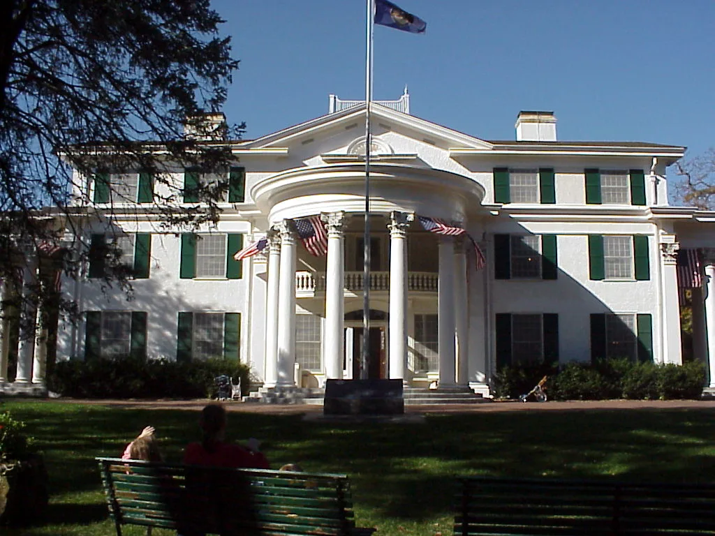 White columned mansion with a lawn and benches in front under a blue sky