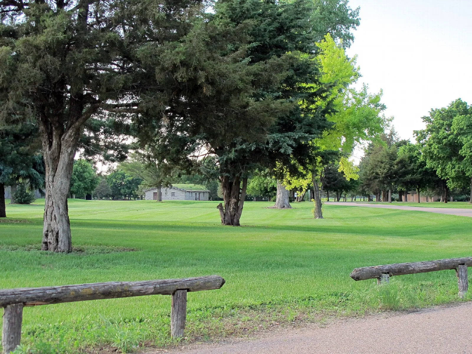 Grassy parade ground with shade trees and a log rail beside a paved path