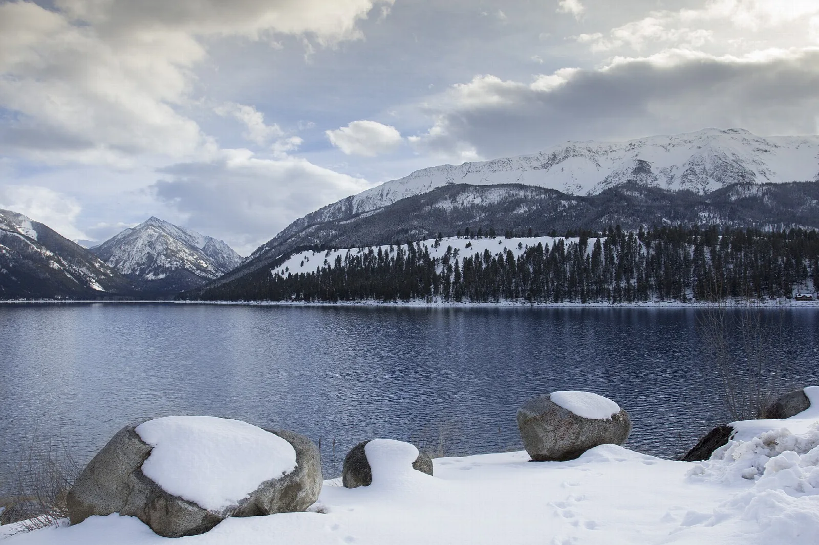 Snowy mountains above a dark lake with snow-capped boulders on the shore