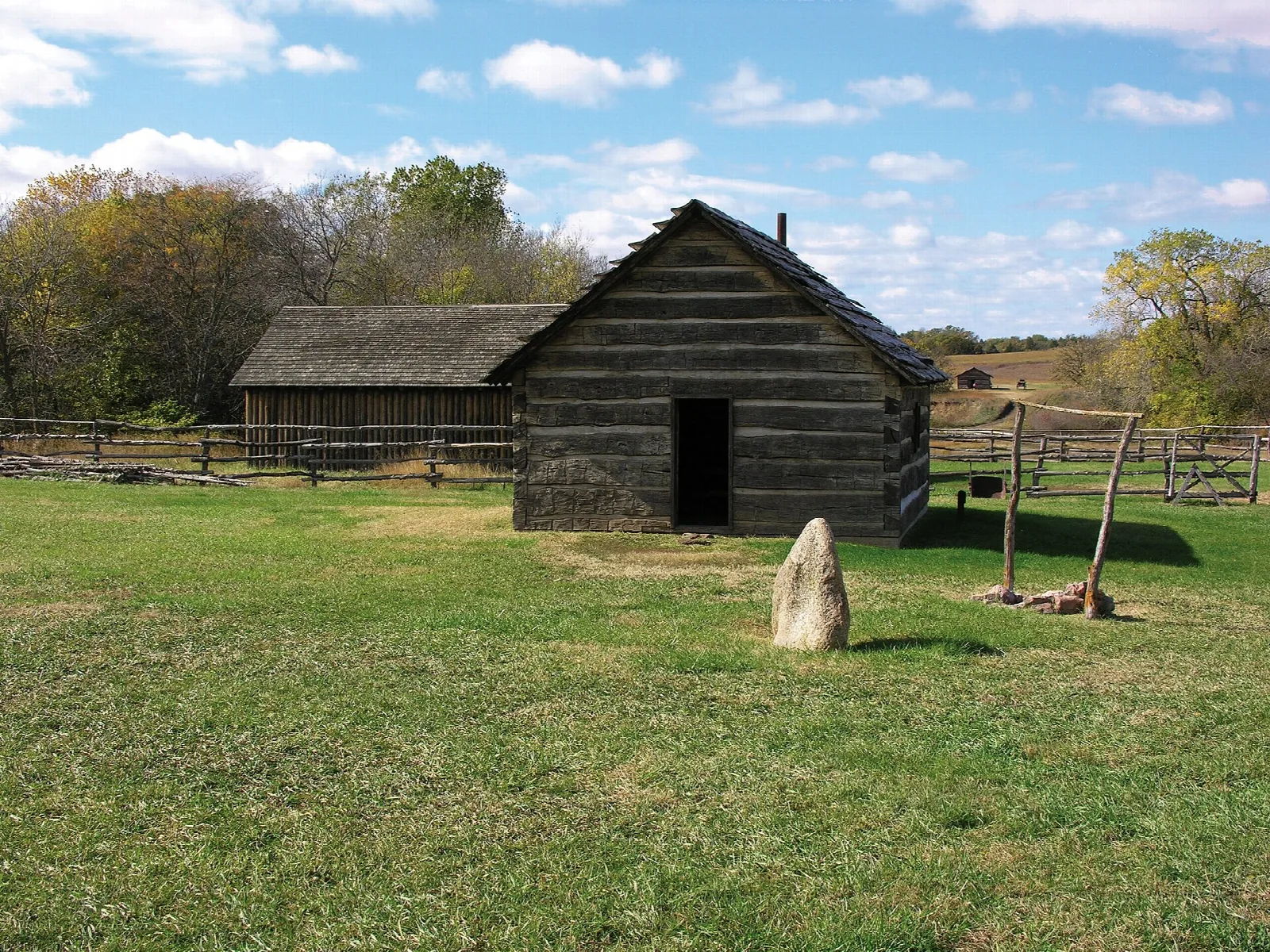 Log cabin with a stone marker and a split-rail fence on a green lawn