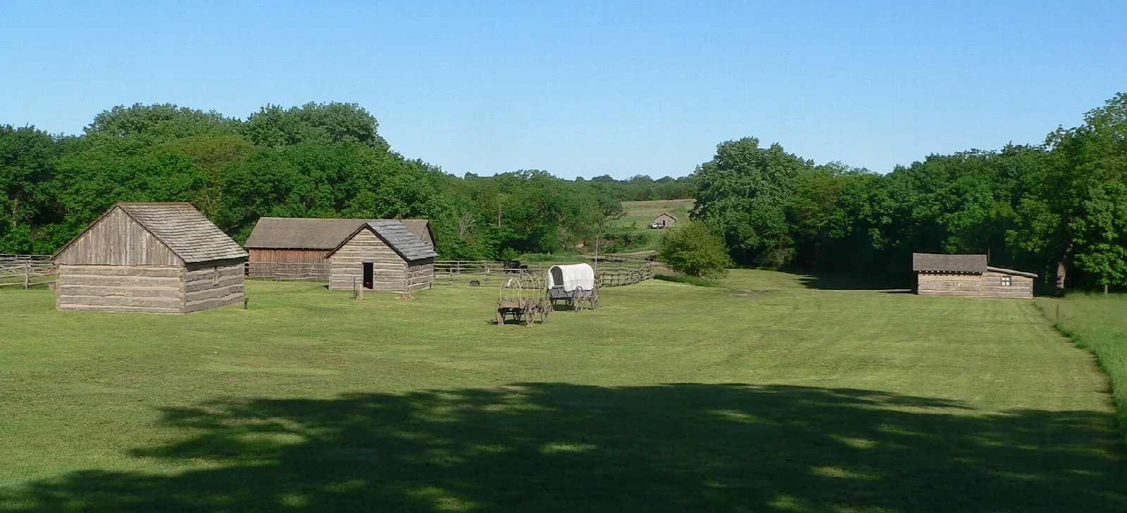 Reconstructed log buildings and a wagon across a wide mown field edged with trees