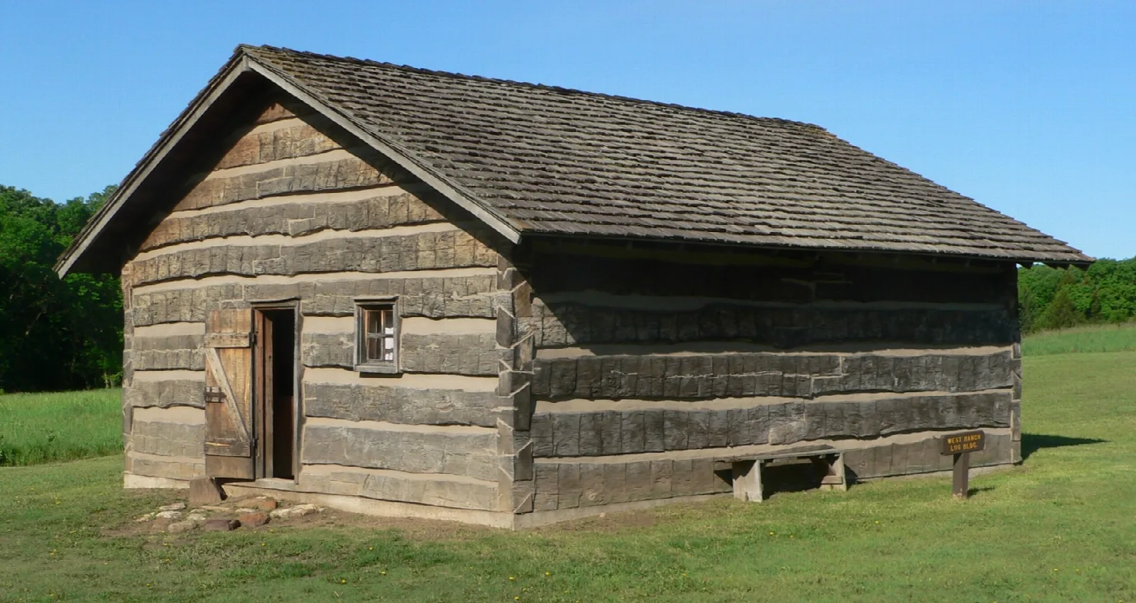 Weathered log building with a shingled roof and a small door on a lawn