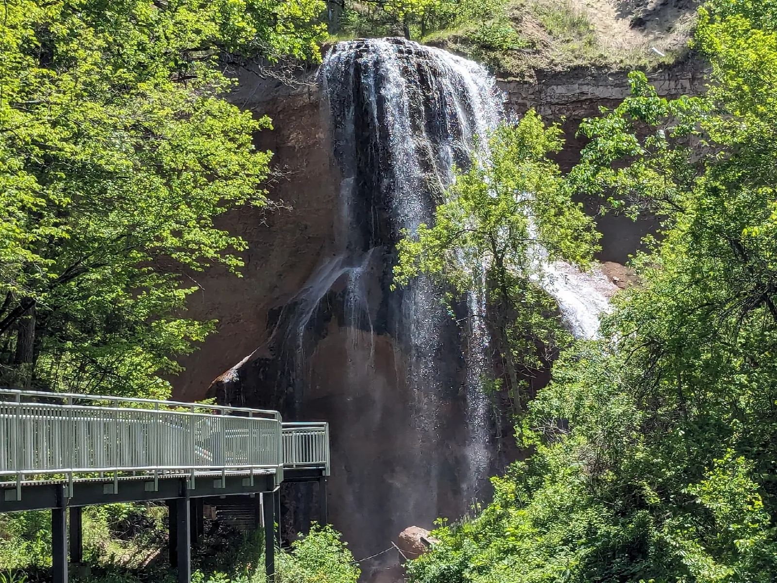 Waterfall dropping down a rock face among green trees, with a walkway at left