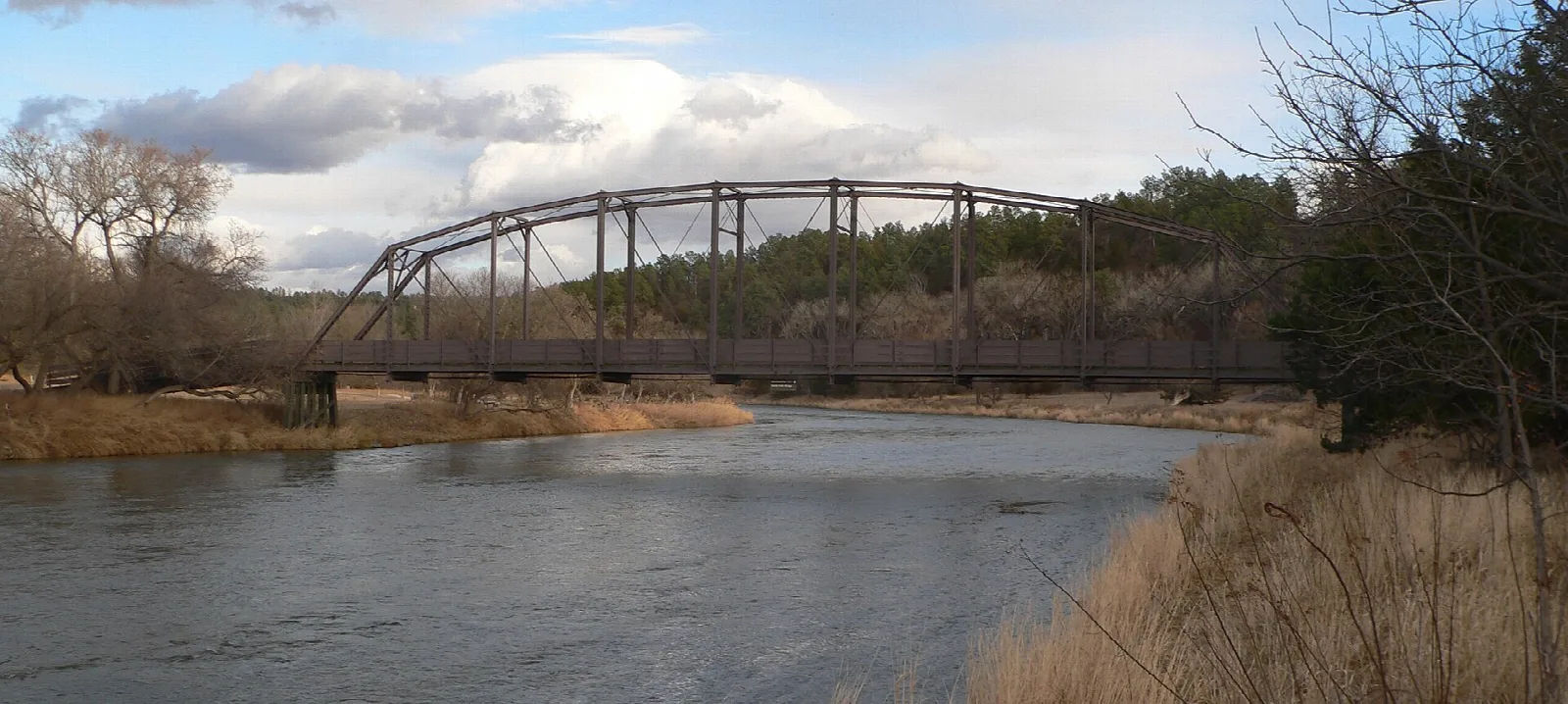 Steel arched footbridge crossing a calm river with bare trees on the bank