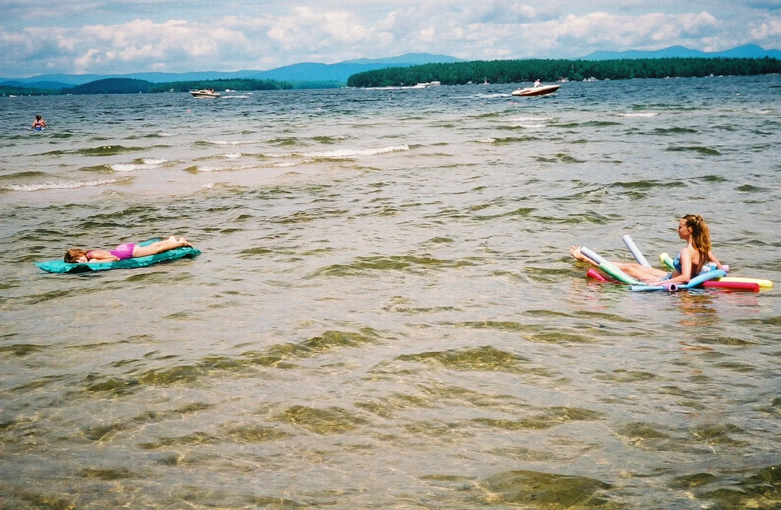Swimmers on floats in shallow lake water with hills and boats in the distance