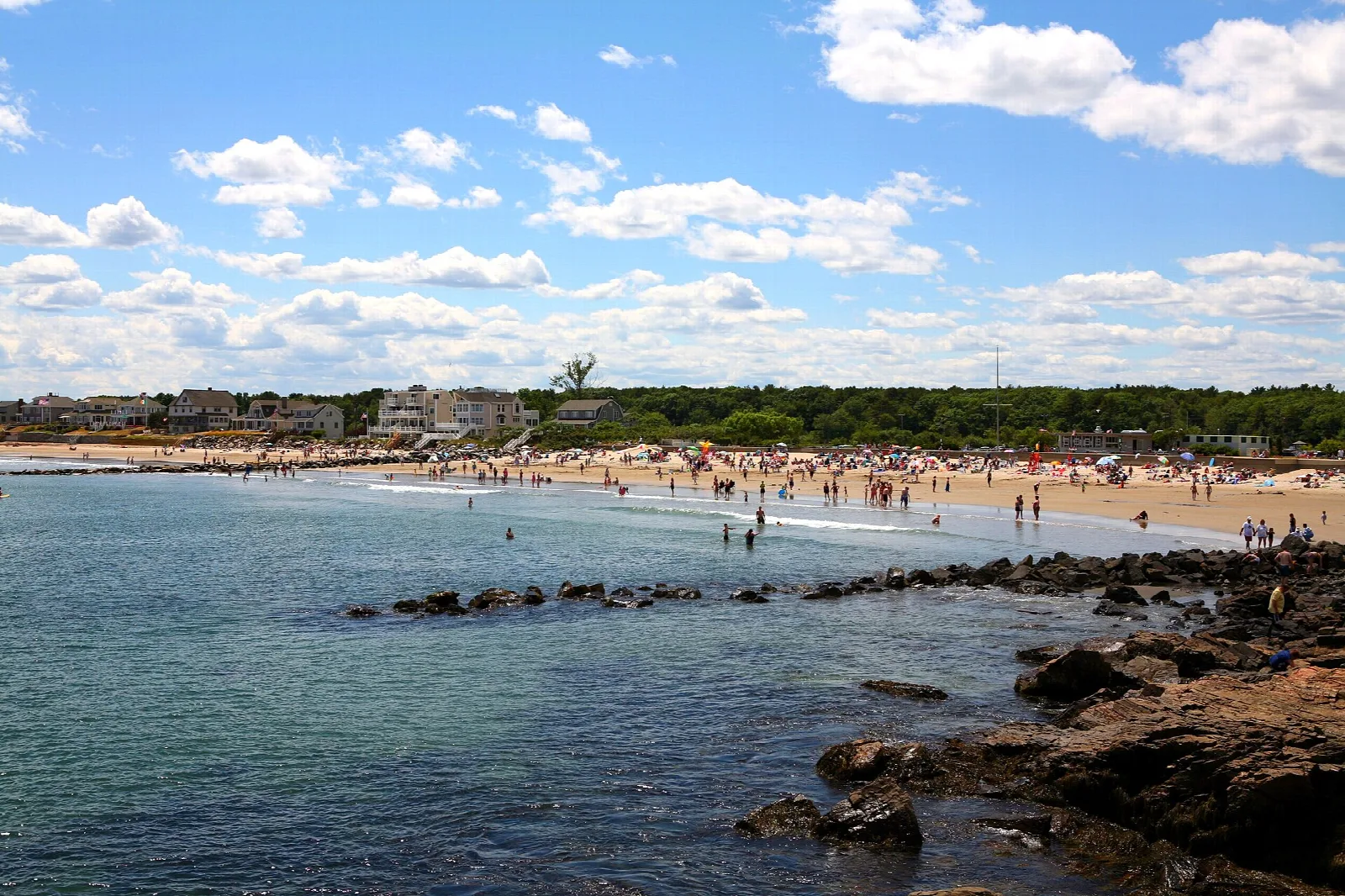 Crowded sandy beach beyond a rocky shoreline under a bright cloudy sky