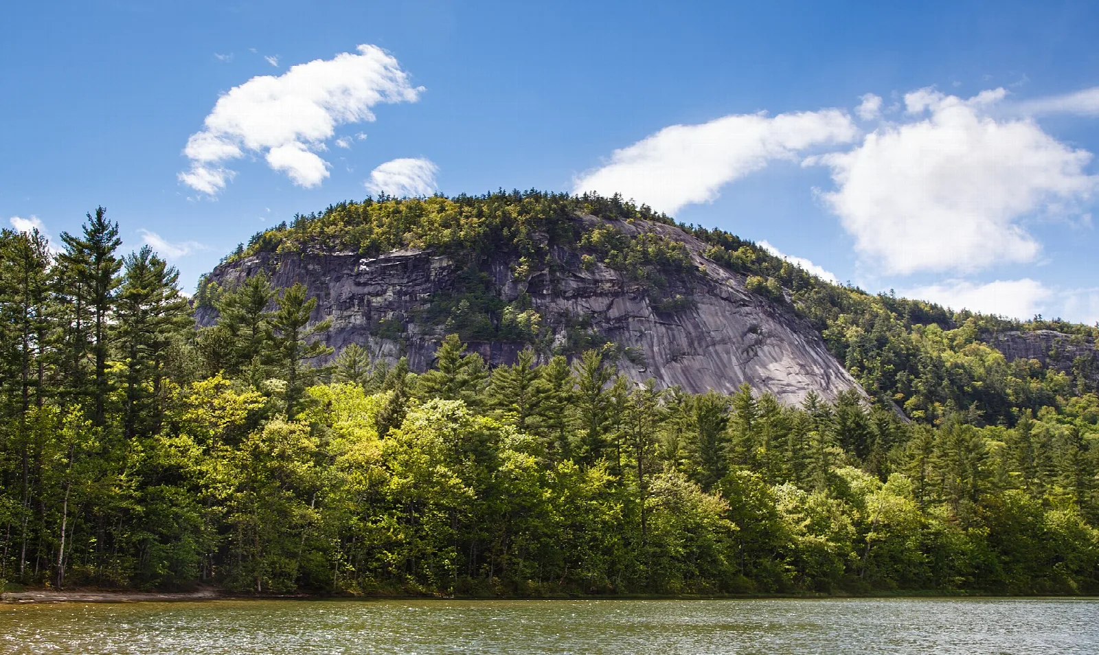 Rounded granite ledge above green trees under a blue sky with clouds