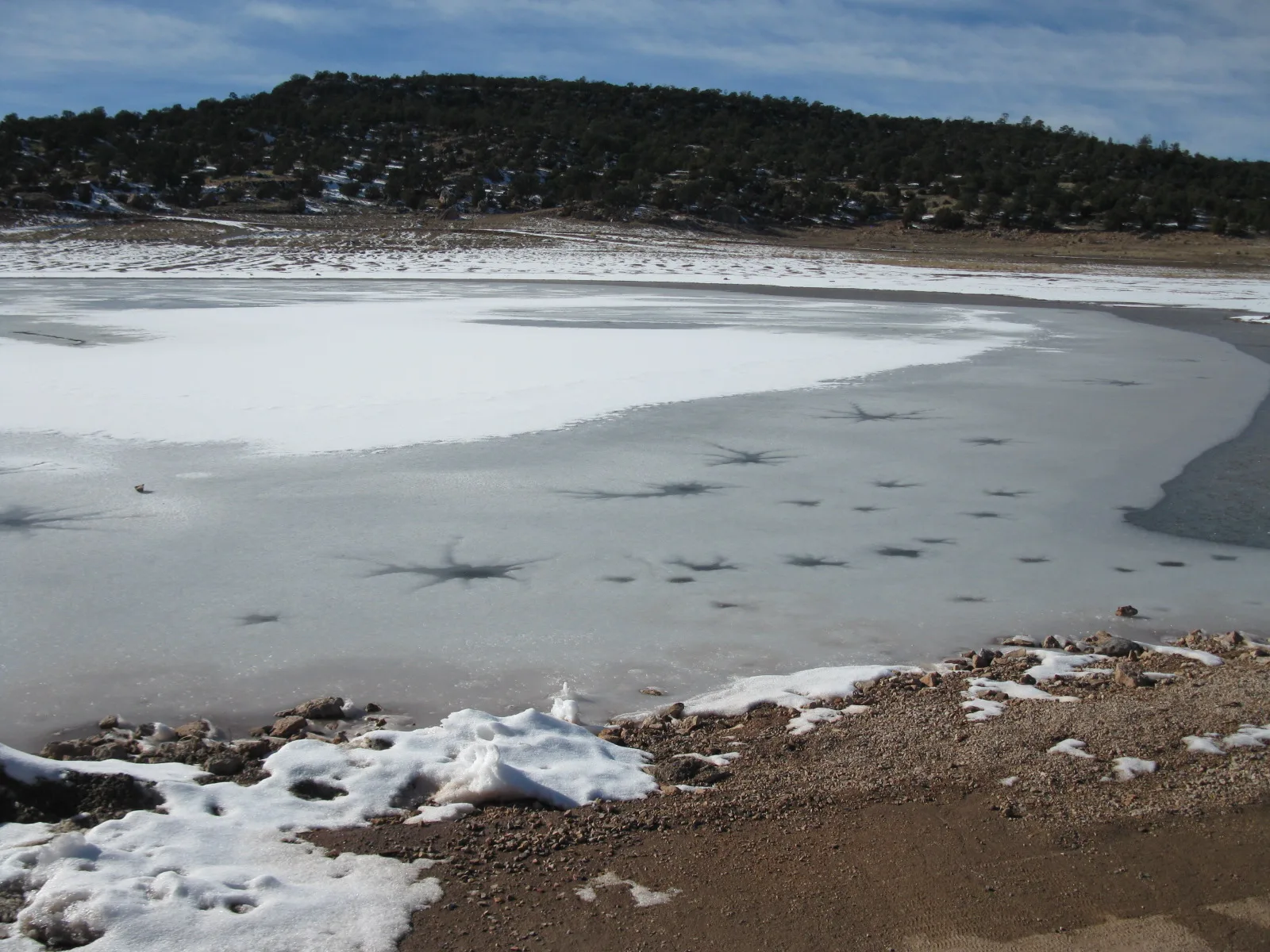 Snow and ice along the shore of a lake, with a forested ridge beyond