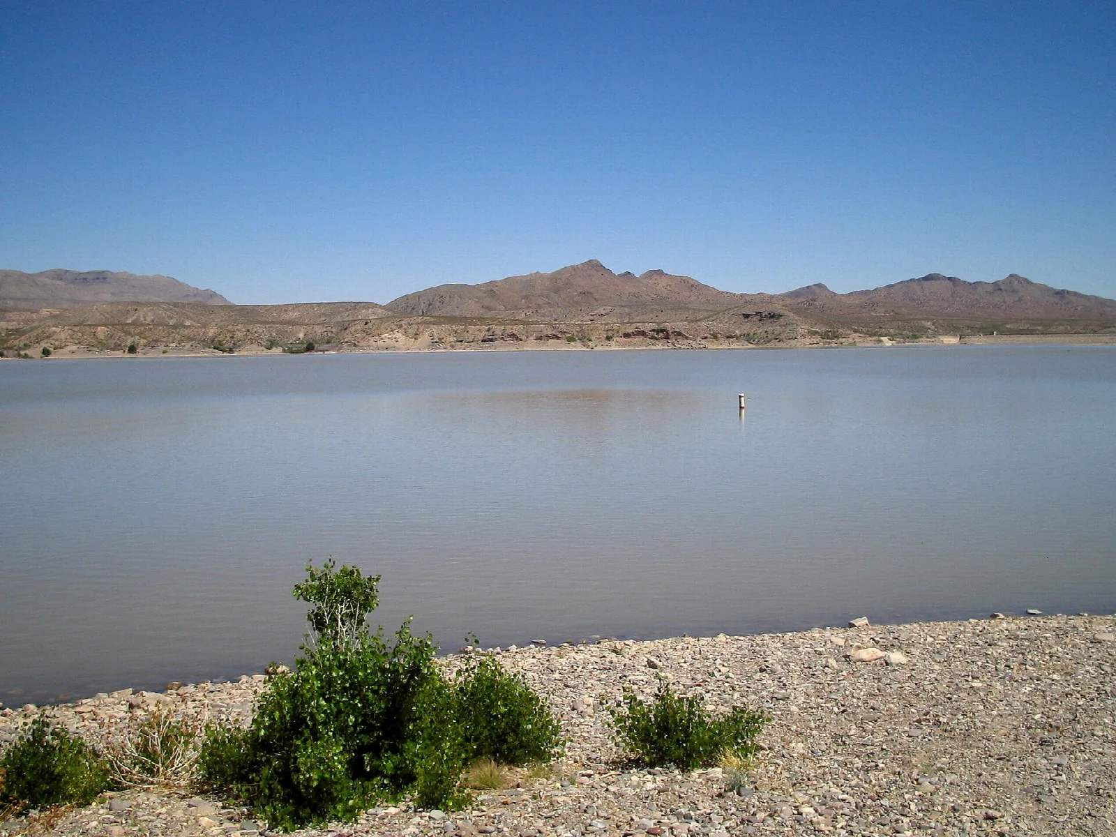 Calm lake with a gravel shore and dry brown mountains under a clear blue sky