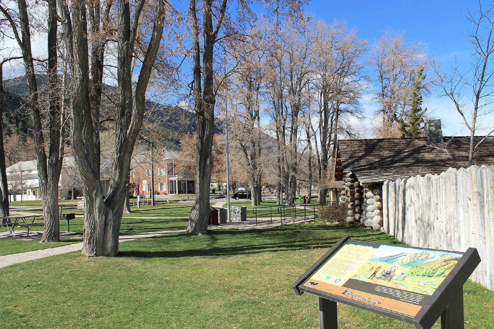 Green lawn with a log stockade fence, an information sign and cottonwood trees