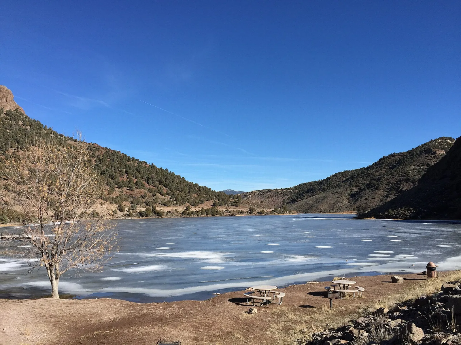 Partly frozen reservoir below tree-covered hills and a blue sky