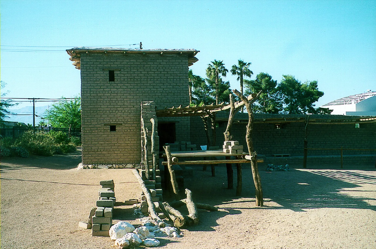Adobe brick tower and wooden framework in a dusty yard with palm trees