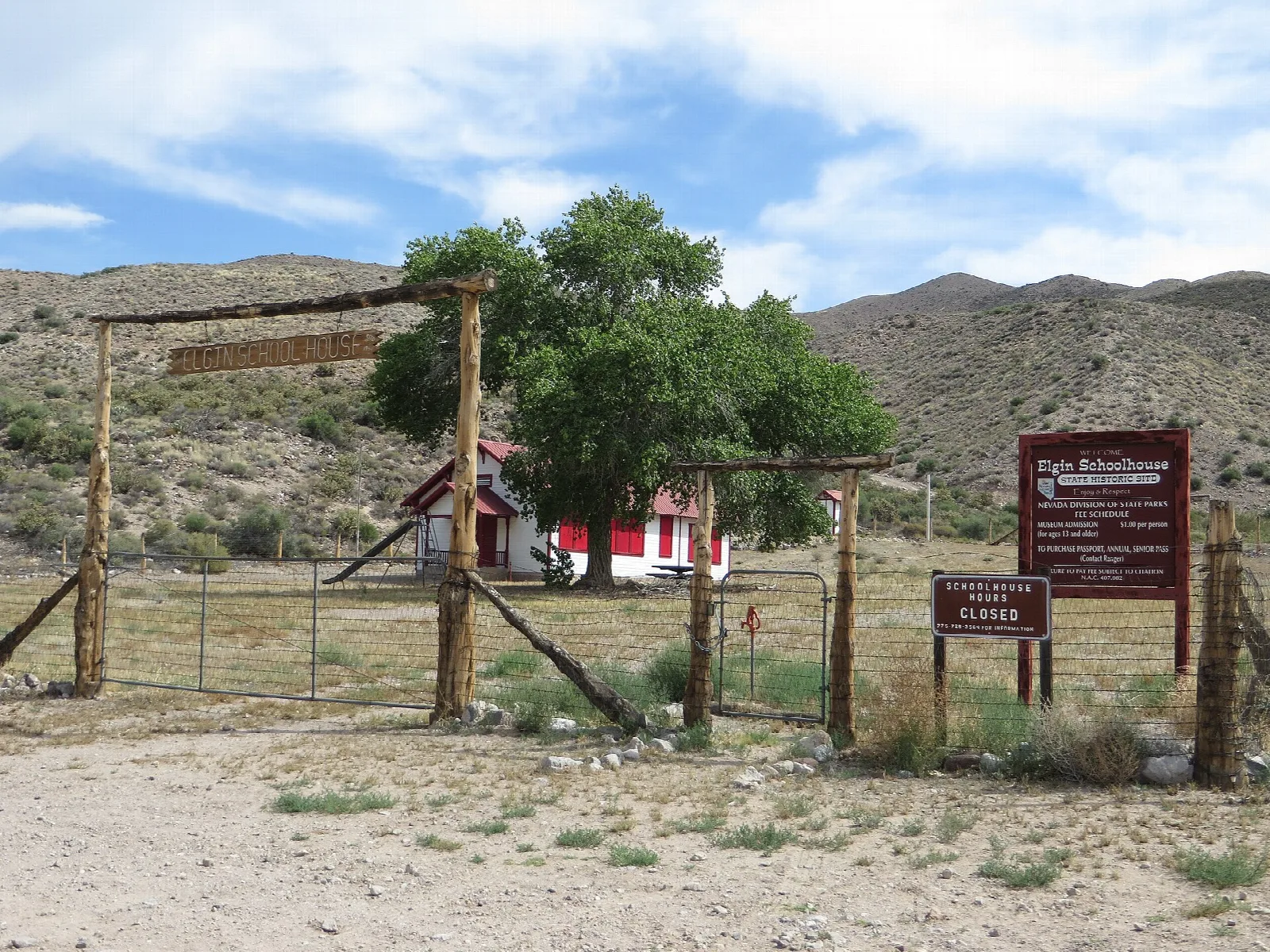 Red and white schoolhouse behind a wooden gate and fence under a cottonwood tree