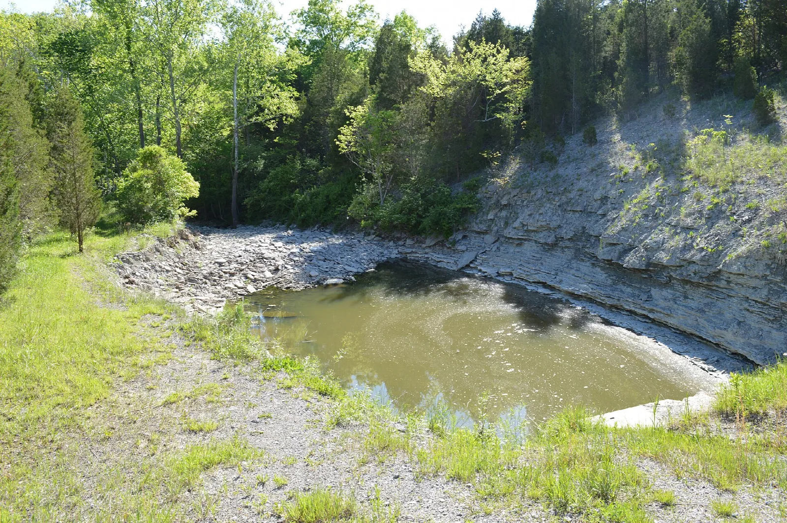 Layered rock wall curving around a shallow pond with a grassy shore