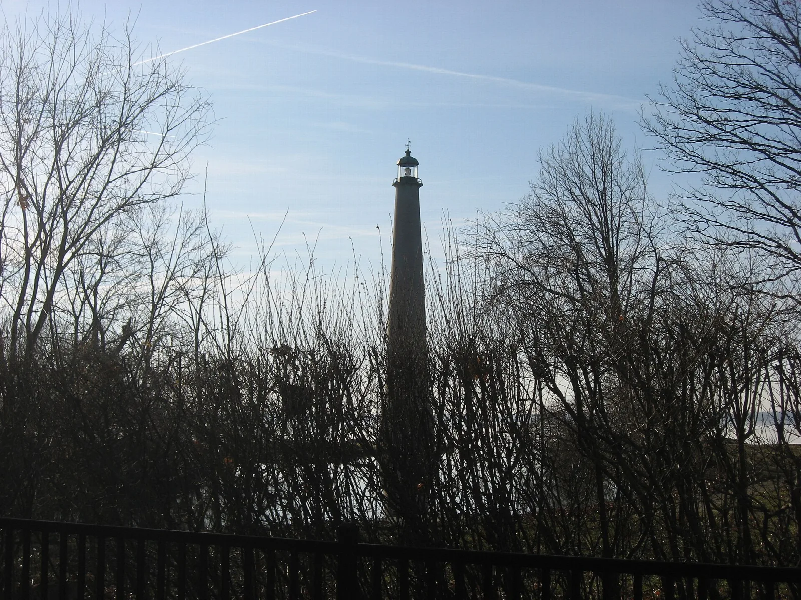 Tall brick lighthouse tower behind bare trees and a railing