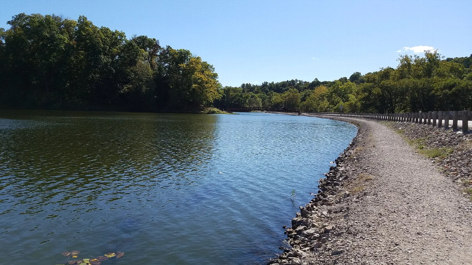 Lake with a gravel path along the shore and wooded hills beyond