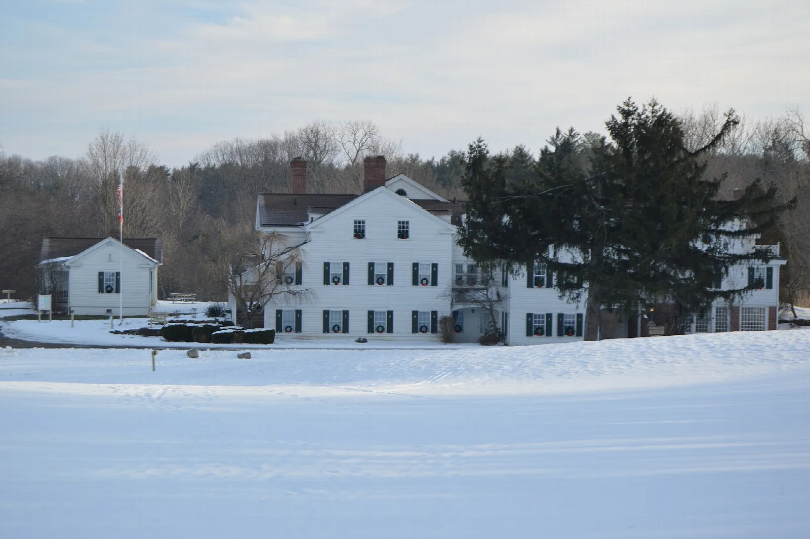 White historic manor house with outbuildings beside a snow-covered field and bare trees