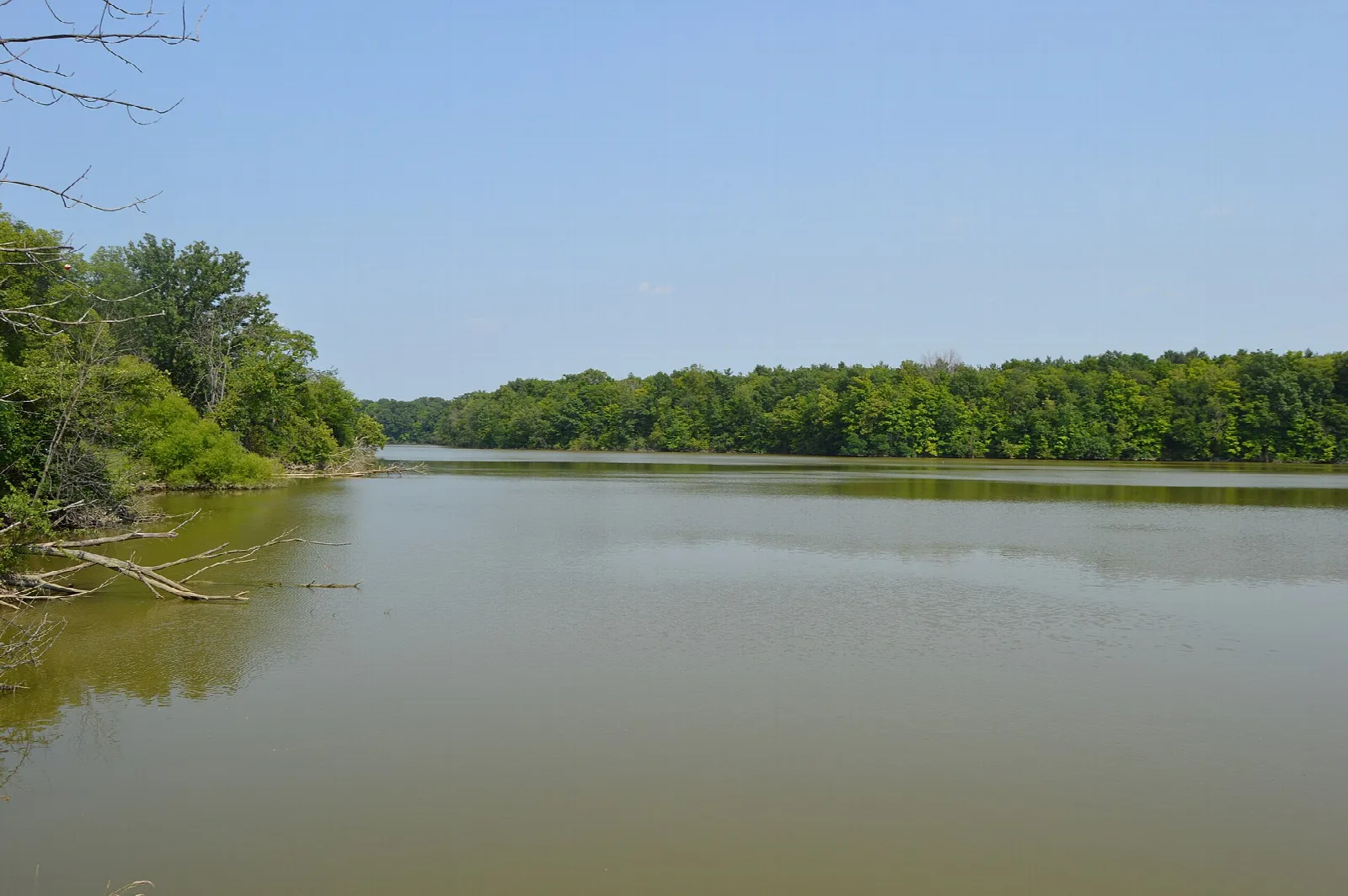 Calm lake seen from a spillway with tree-lined shores under a hazy sky
