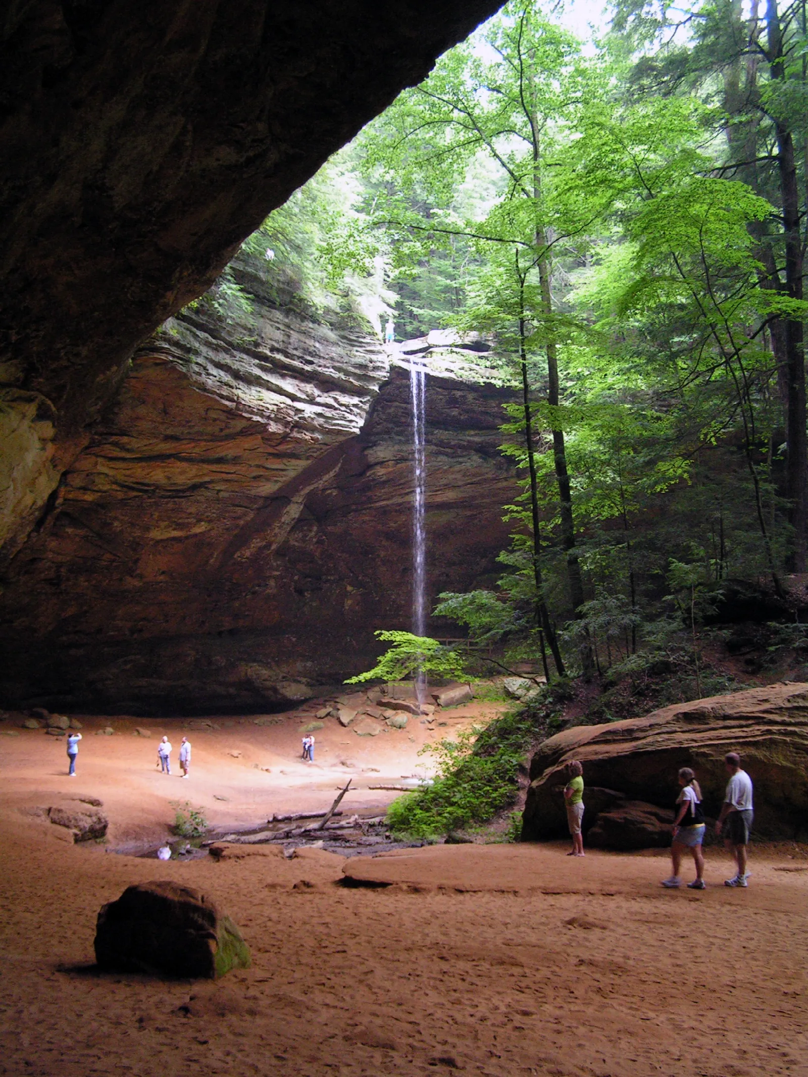 Tall recess cave with a waterfall and a few visitors on sandy floor