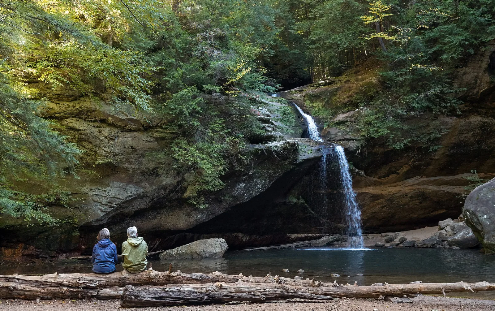 Waterfall dropping into a pool under a rock overhang with two people sitting on a log