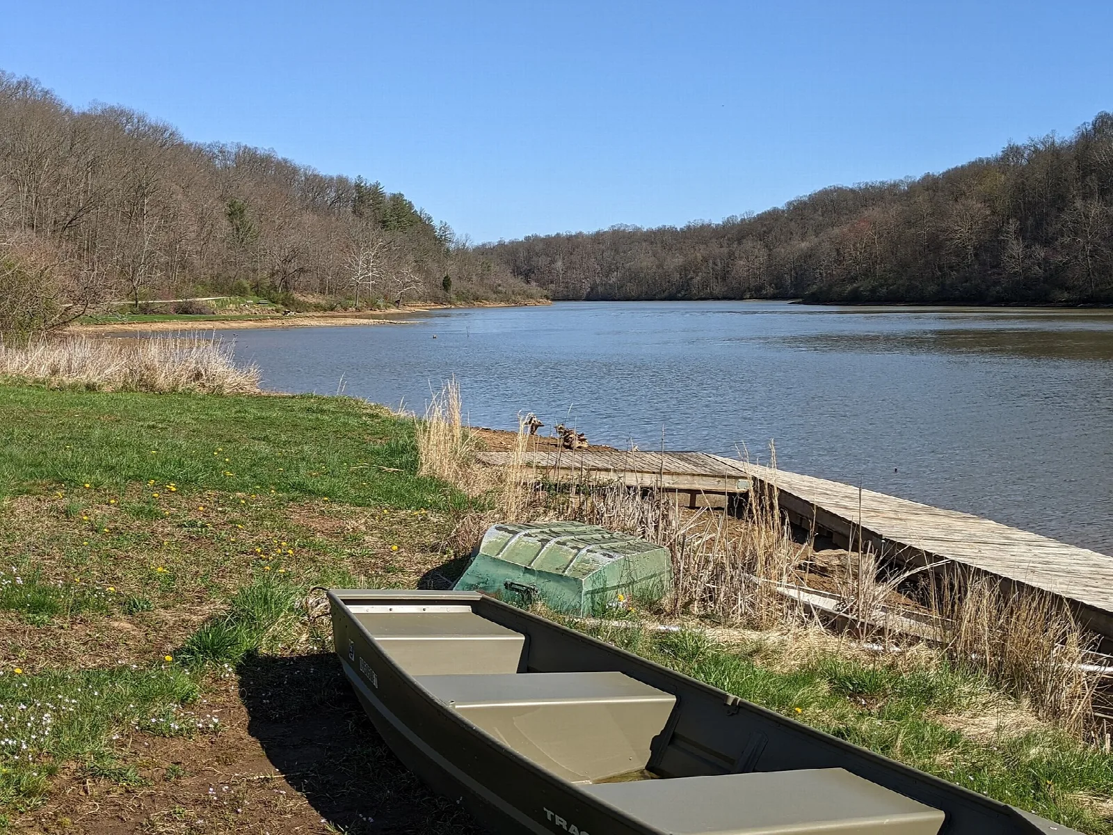 Small boat pulled up on a grassy lake shore with a dock and wooded hills