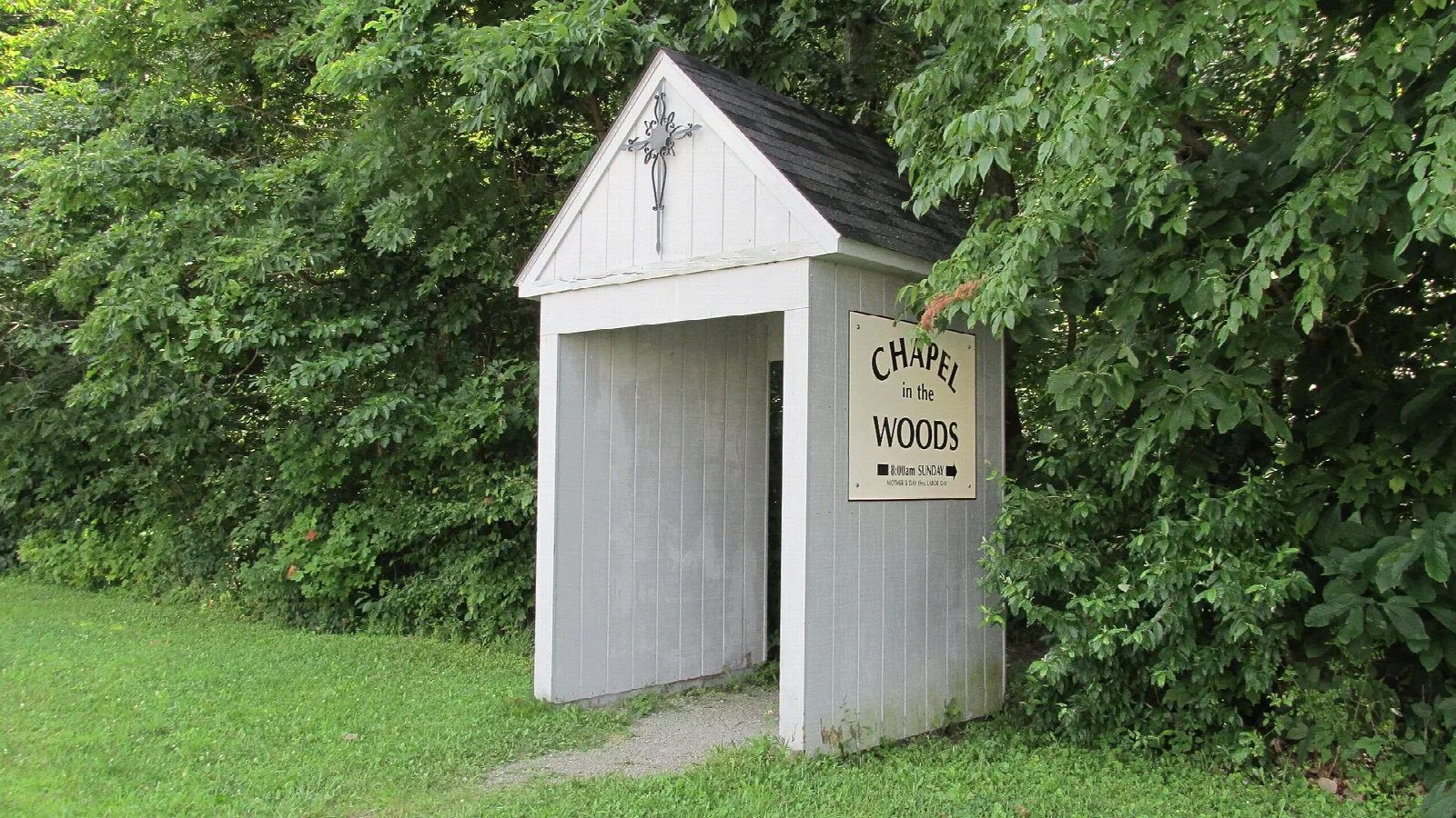 Small white open-sided chapel with a sign among green trees
