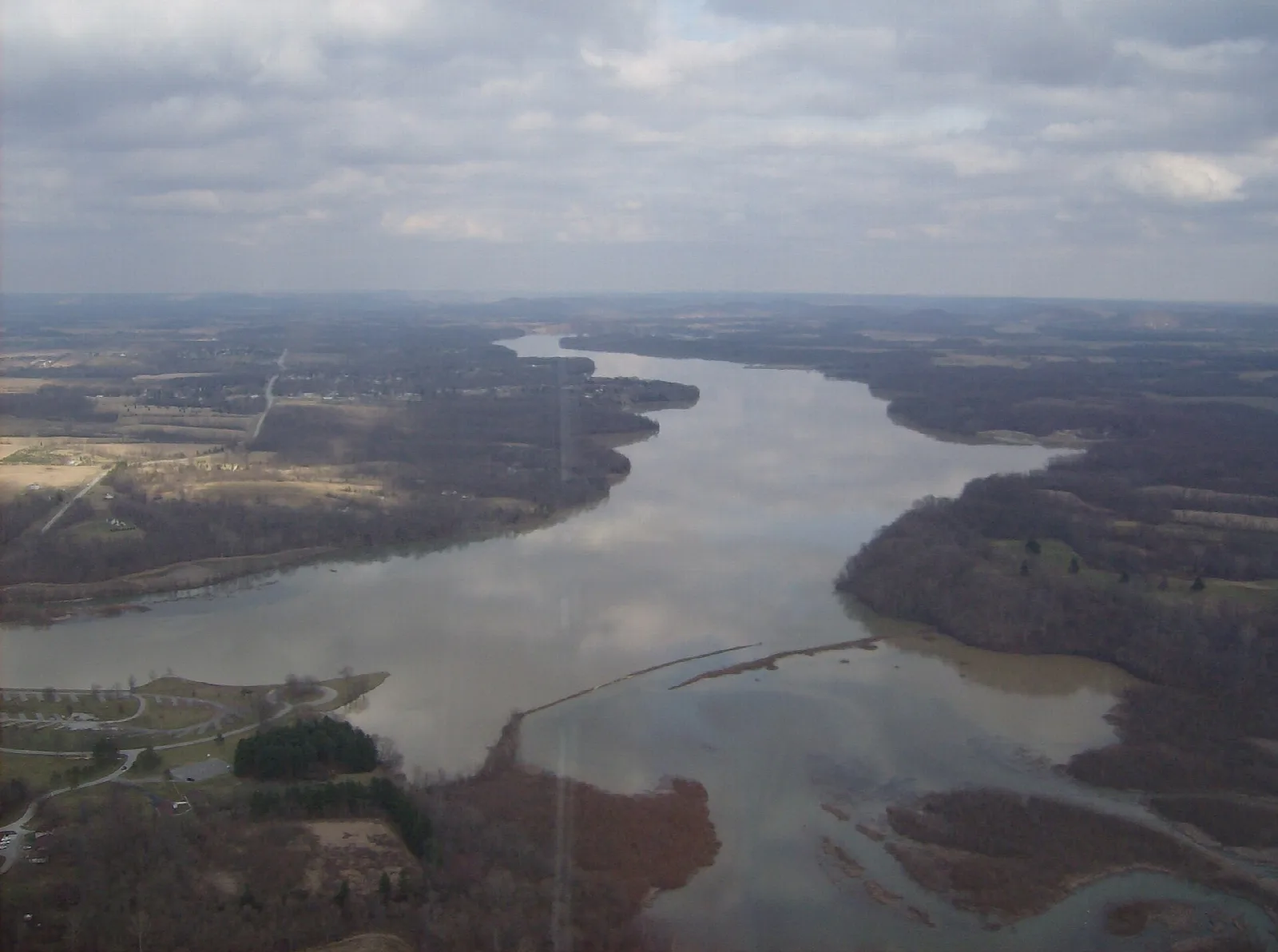 Aerial view of a wide winding lake with fields and woods along the shore under gray clouds
