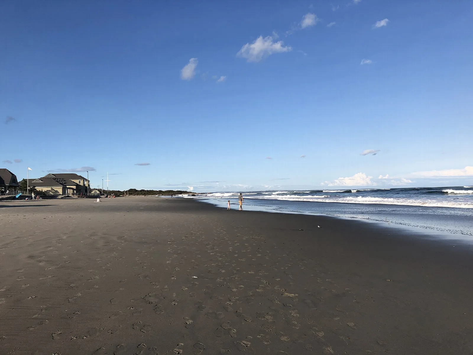Wide dark-sand beach with surf breaking and beach buildings in the distance