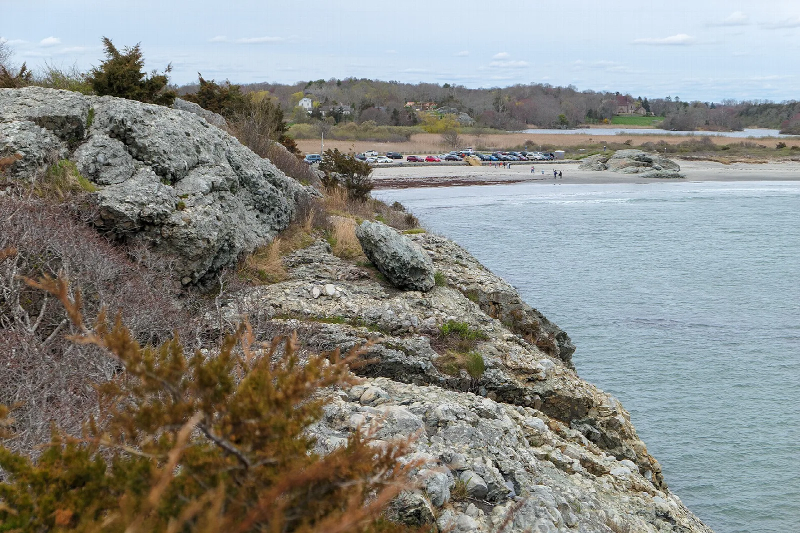 Rocky cliff edge above water with a beach and parked cars in the distance