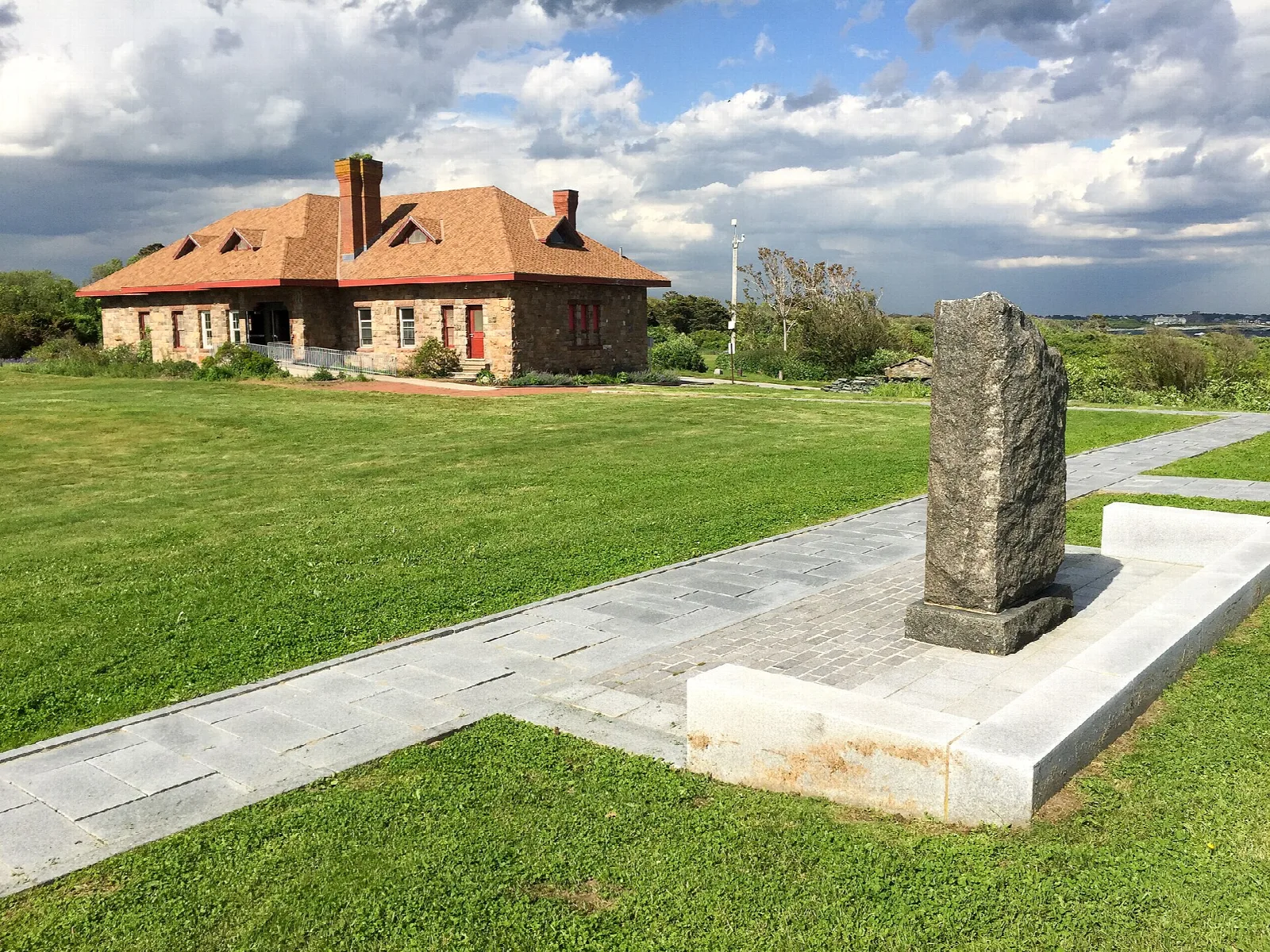 Stone building with a red tile roof beyond a lawn and a stone marker