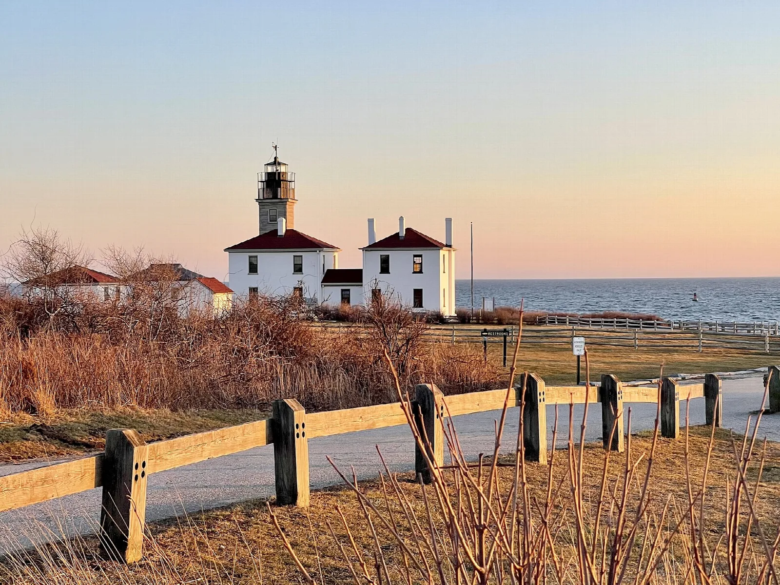 Lighthouse and houses at sunset behind a wooden fence beside a road